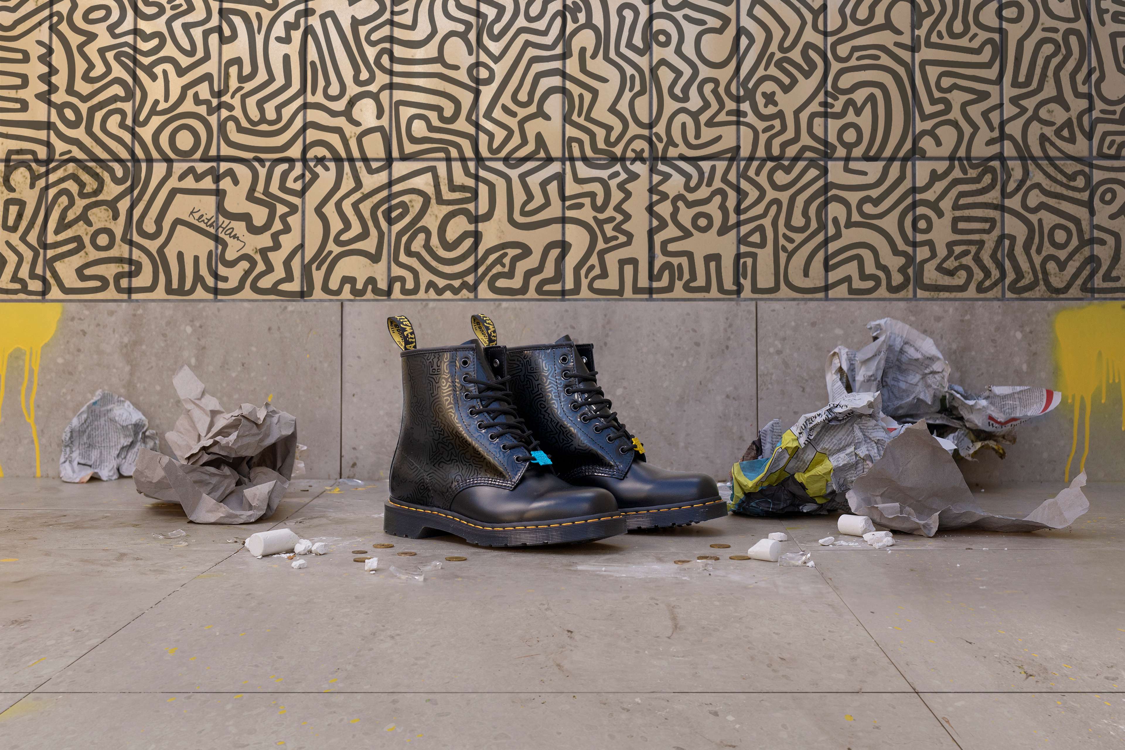 Une photographie d'une paire de bottes issues de la collaboration entre Dr. Martens et Keith Haring, posée devant un arrière-plan présentant l'une des fresques murales emblématiques de l'artiste.