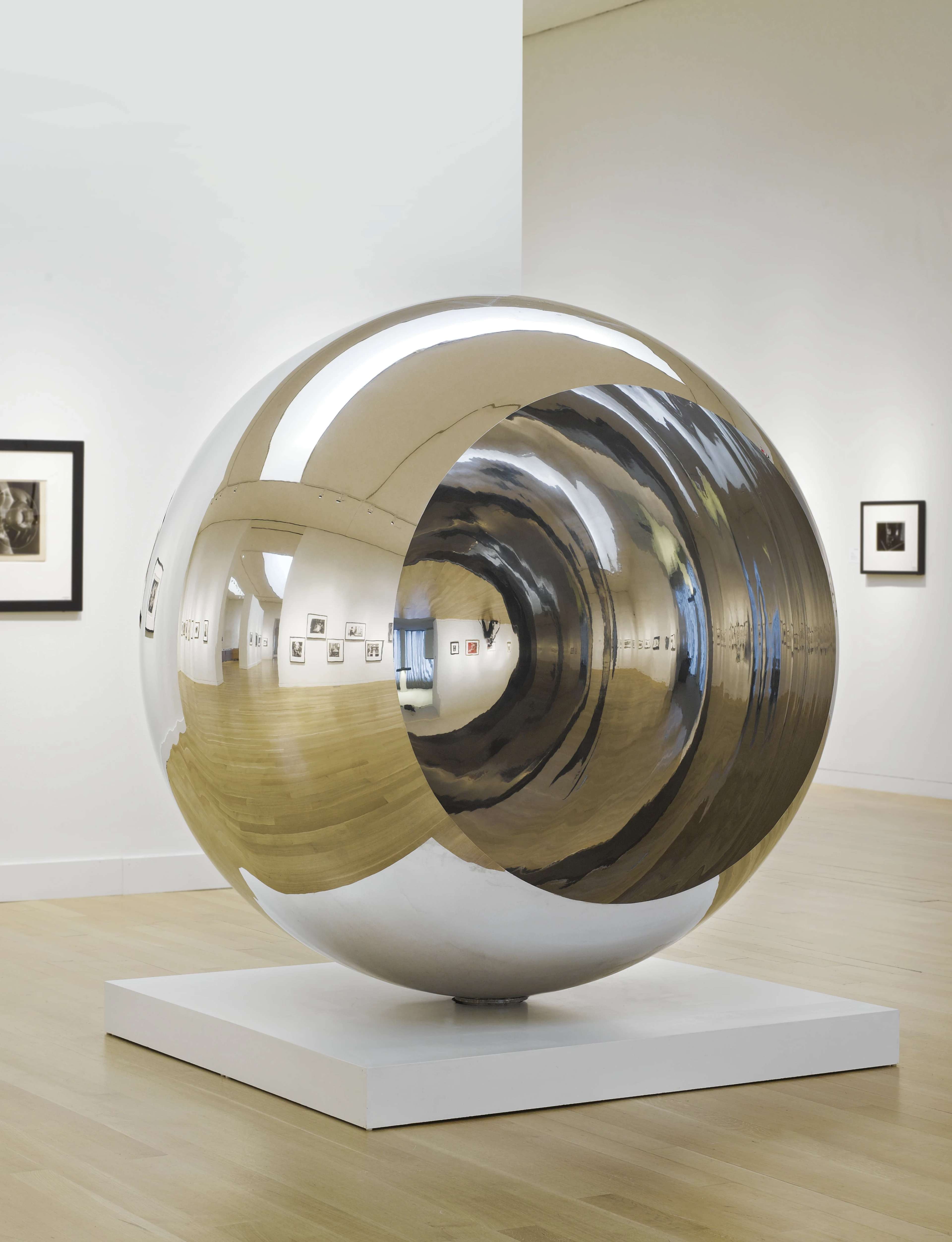 A reflective hollow mirror sphere, balanced on a flat, square, white plinth. The sphere is open on both sides, allowing the viewer to look through it and see inside it. Its outside and inside reflect different perspectives on the gallery space around it.