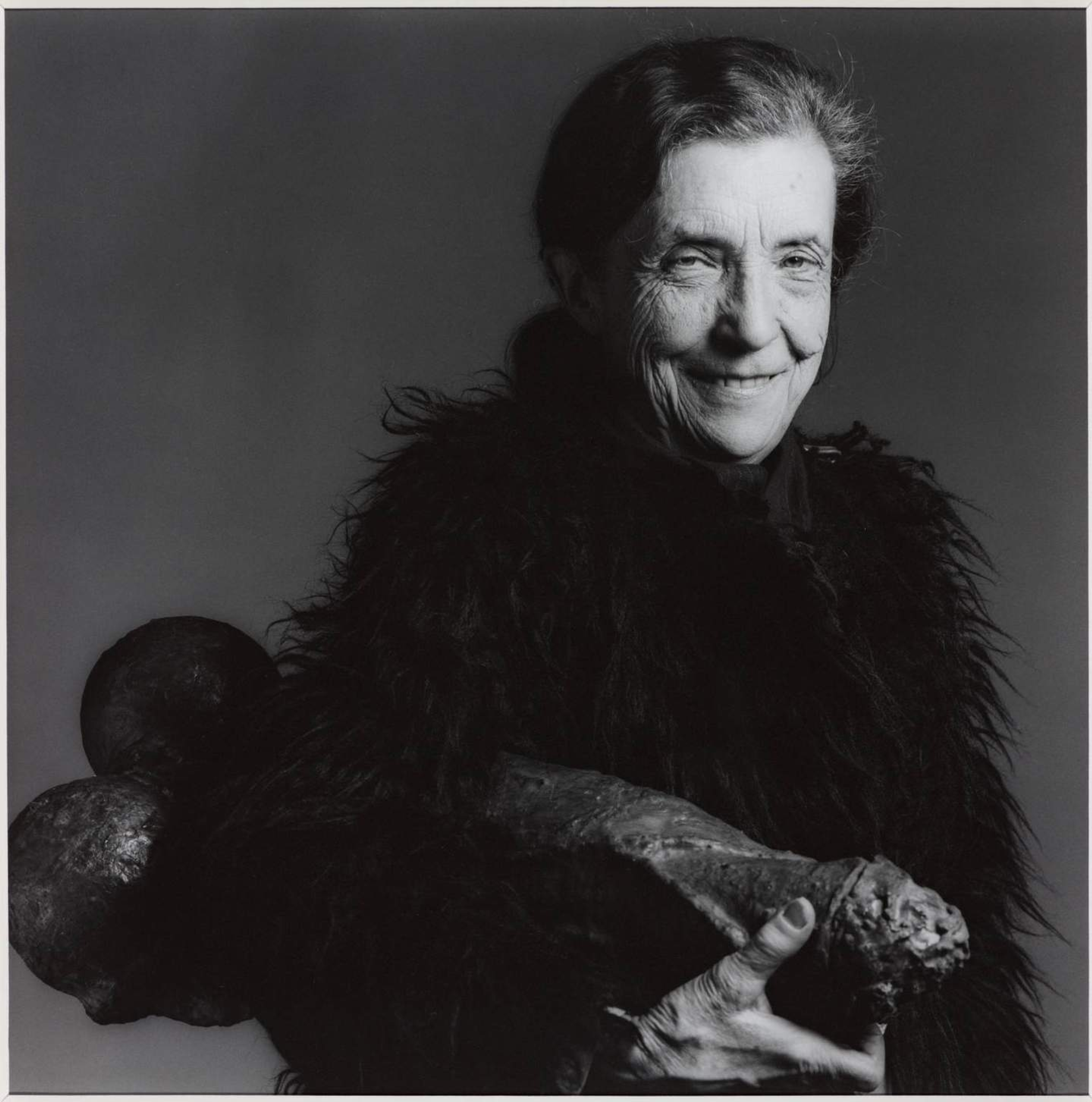 Photographie en noir et blanc de l'artiste Louise Bourgeois souriant, tenant un objet de forme phallique sous son bras.
