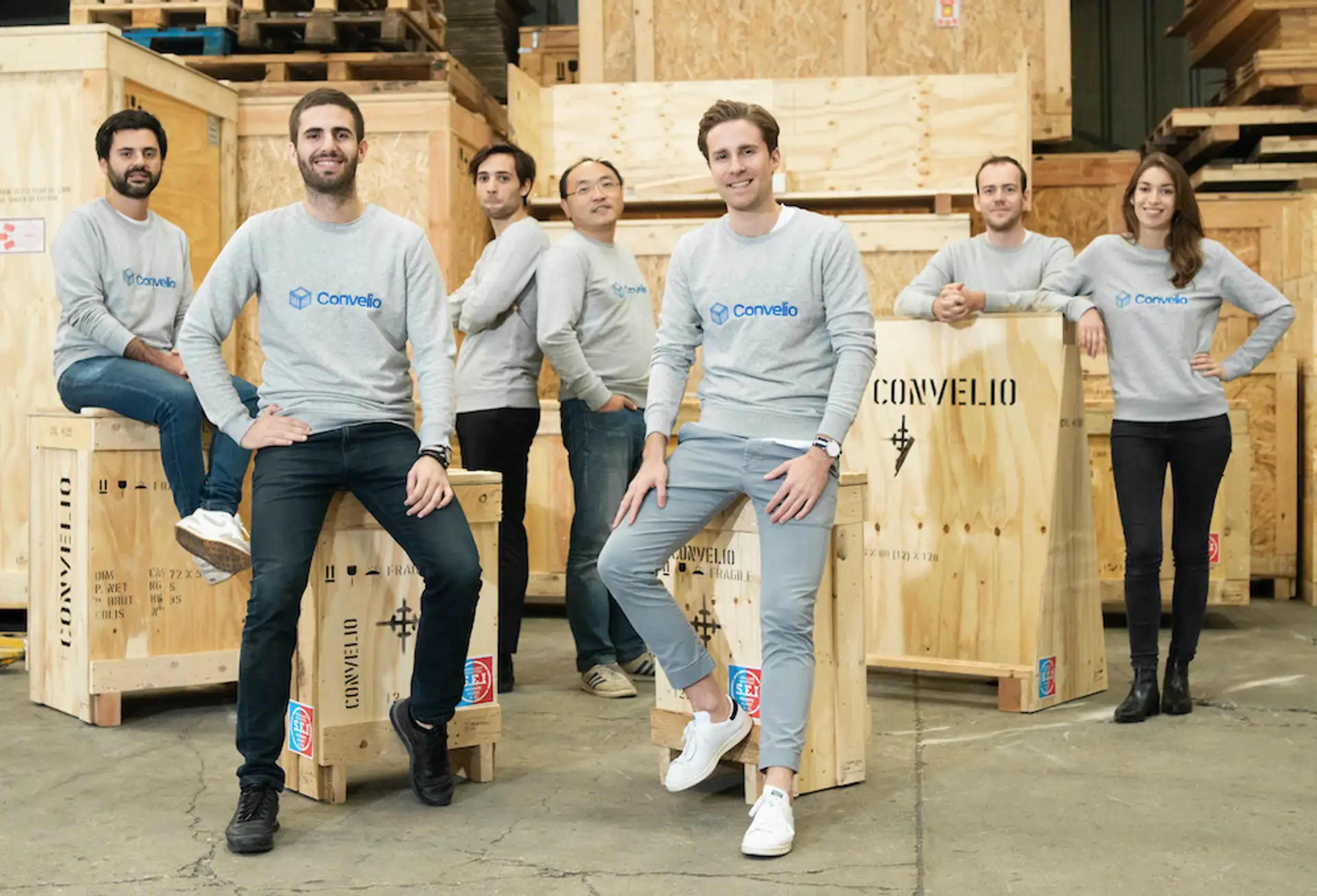 Une photo de l'équipe Convelio, tous portant des pulls gris assortis, appuyée contre des caisses de rangement en bois pour œuvres d'art.