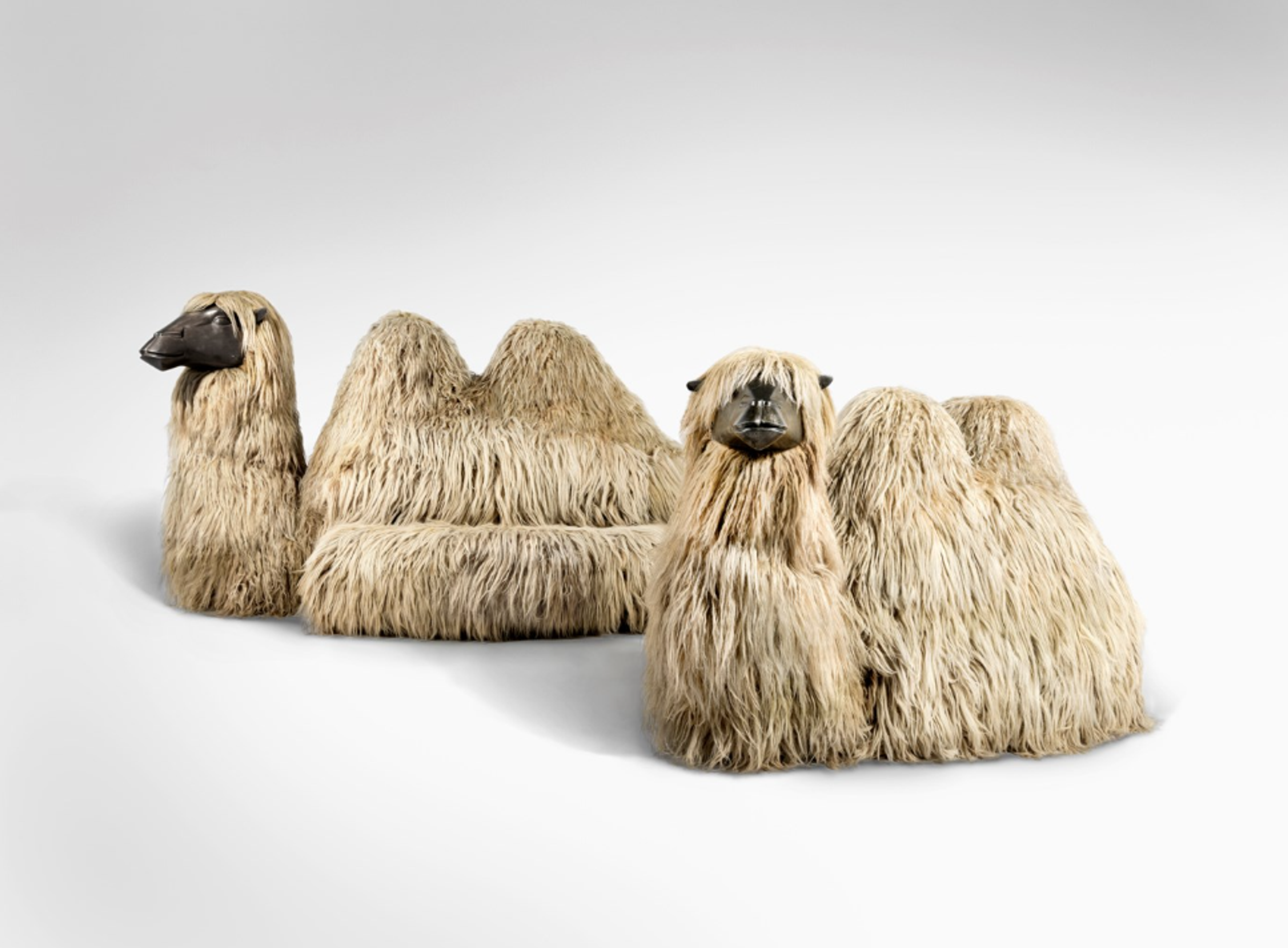 A mixed-media sculpture featuring two stylised sitting camels with woolly backs and bronze faces. Each has an extra section on one side, allowing them to be used as sofas. They have almost comical expressions.