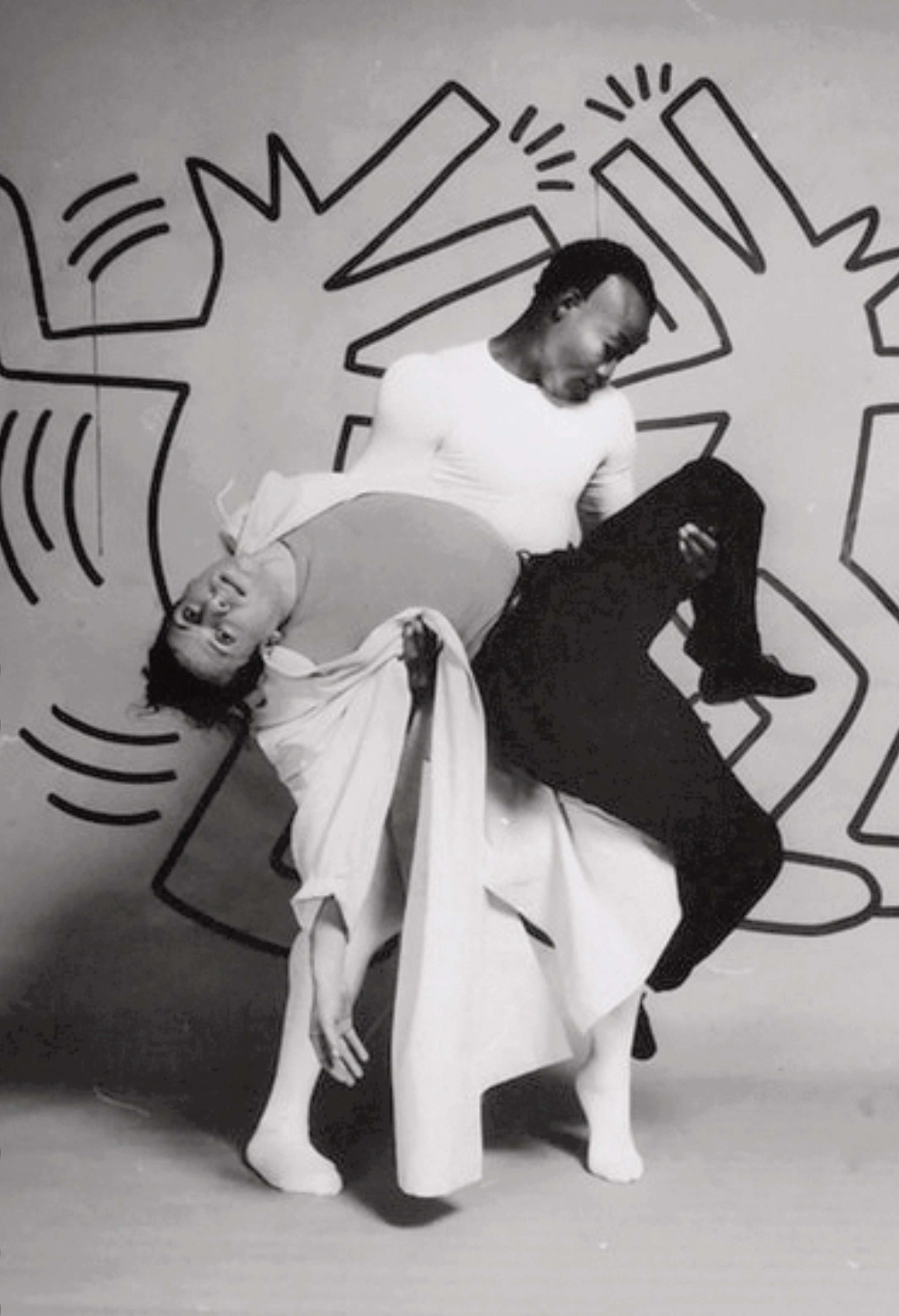 Photographie noir et blanc du décor de Secret Pastures. Deux danseurs posent devant les figures en mouvement de Keith Haring.