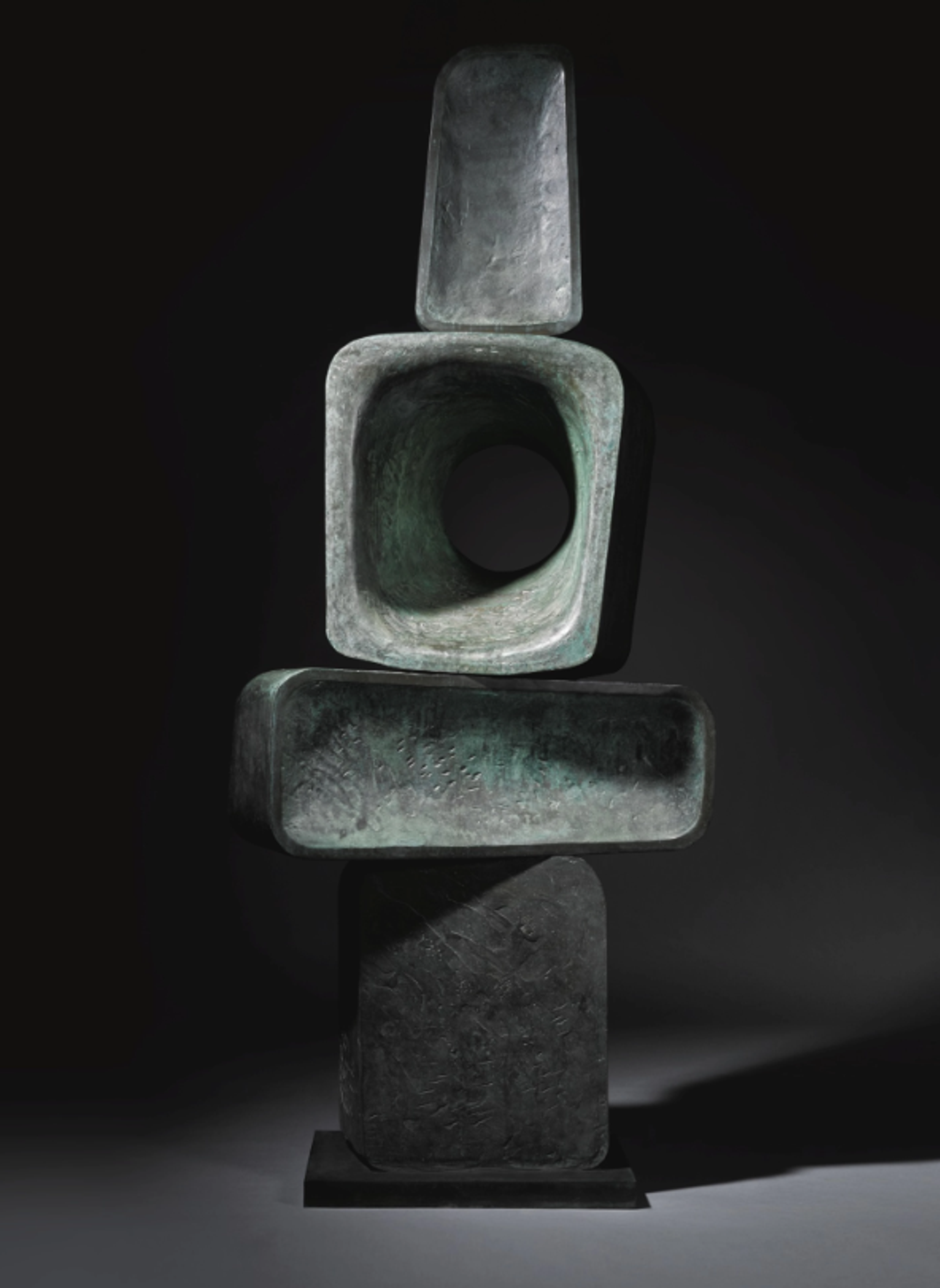 A vertically-oriented abstract bronze sculpture composed of four geometric forms stacked in a totem-like arrangement. The piece features angular shapes including rectangles and squares with hollow openings. The bronze surface has a dark grey patina with subtle green undertones visible in certain areas. The composition, although slightly one-sided, creates a sense of balance between solid forms and negative space.