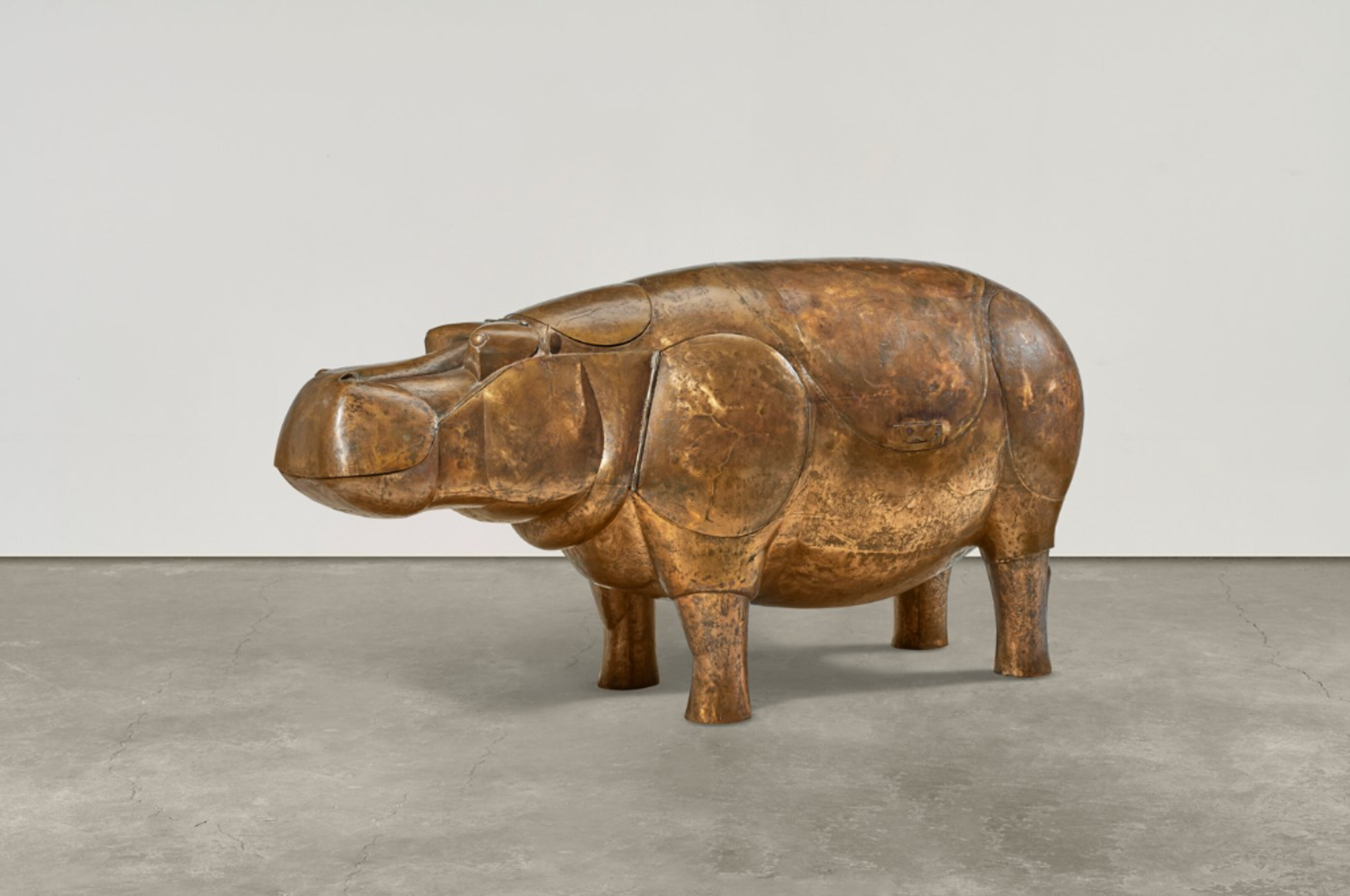 A bronze sculptural bar piece shaped like a hippopotamus. The golden-brown hippo form serves as the functional base, with visible seams and hinges showing where the hippo’s body opens up to reveal the bar within. It has an exaggeratedly smooth and rounded form.