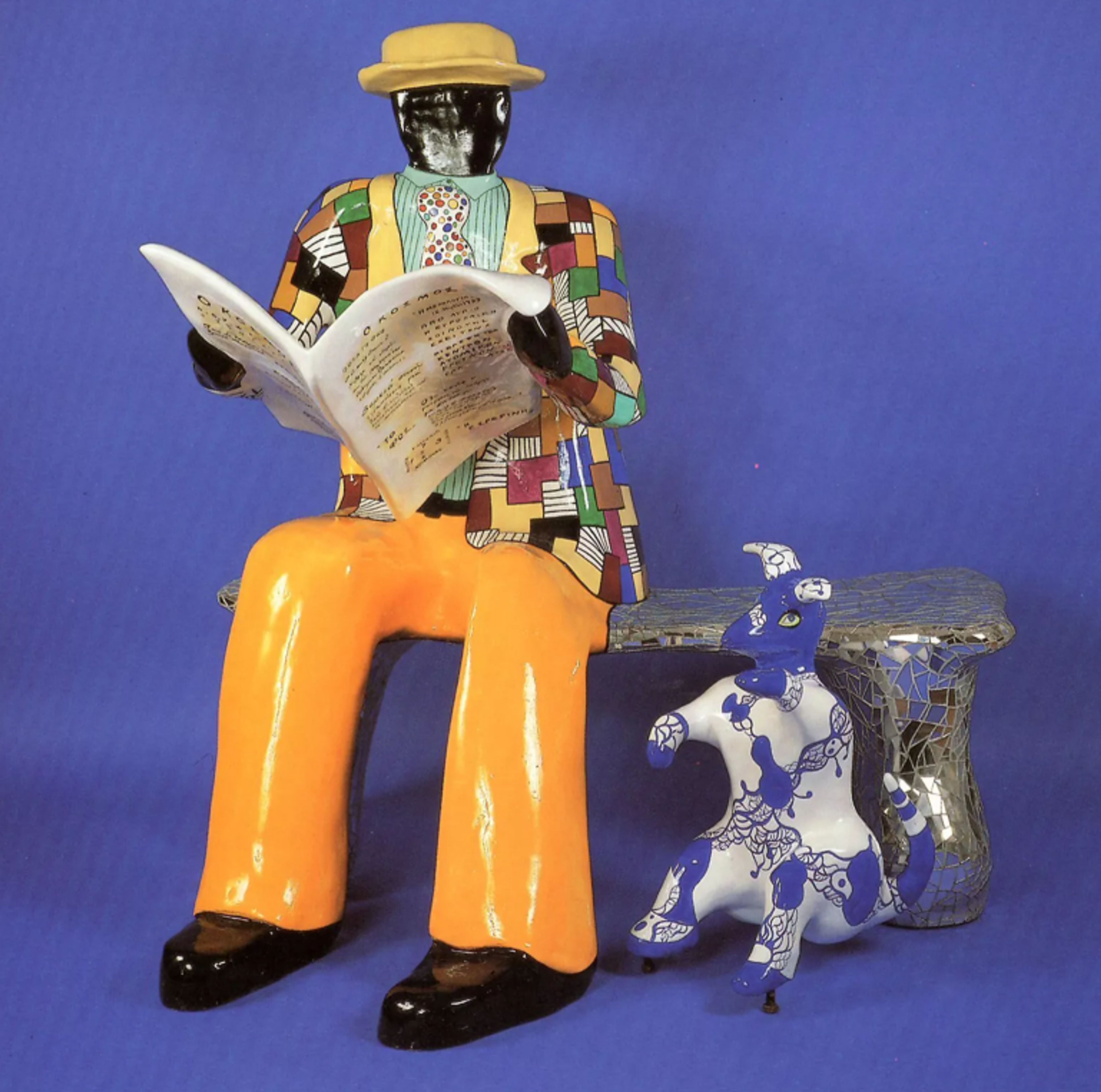 A sculptural scene depicting a seated figure in a vibrant patchwork-patterned outfit and hat, absorbed in reading a Greek newspaper. A small dog in white and blue sits beside him, with his front paws lifted up.