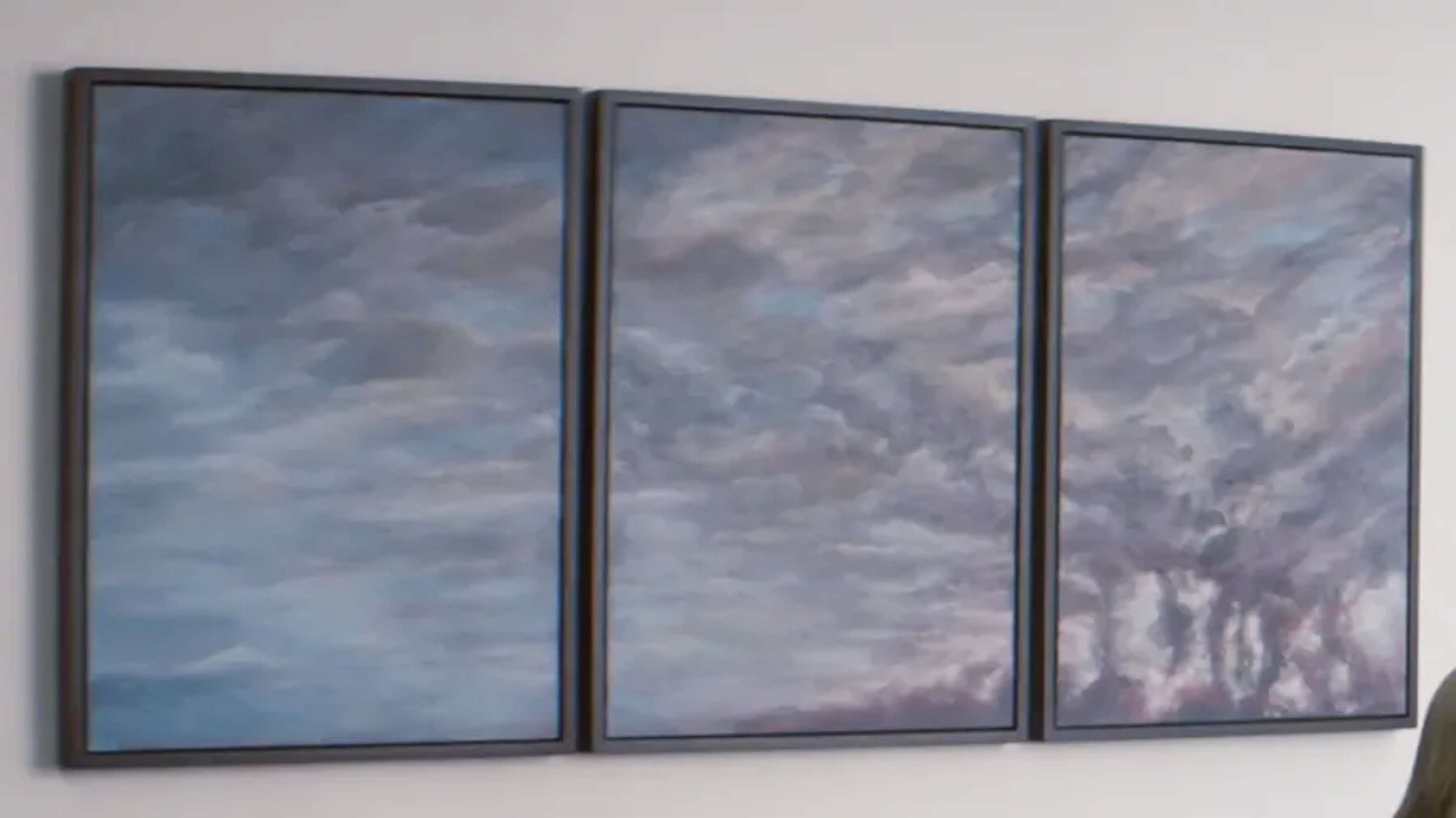 Une peinture en triptyque représentant un ciel sombre et atmosphérique, avec des nuages tourbillonnants dans des tons de bleu, de gris et des touches de violet. L'œuvre en trois panneaux est encadrée de noir et montée sur un mur blanc. Le panneau de droite présente des formations nuageuses plus sombres et dramatiques, avec des traînées évoquant la pluie ou de la fumée s'élevant du bas, ce qui ajoute une impression de mouvement et de profondeur.