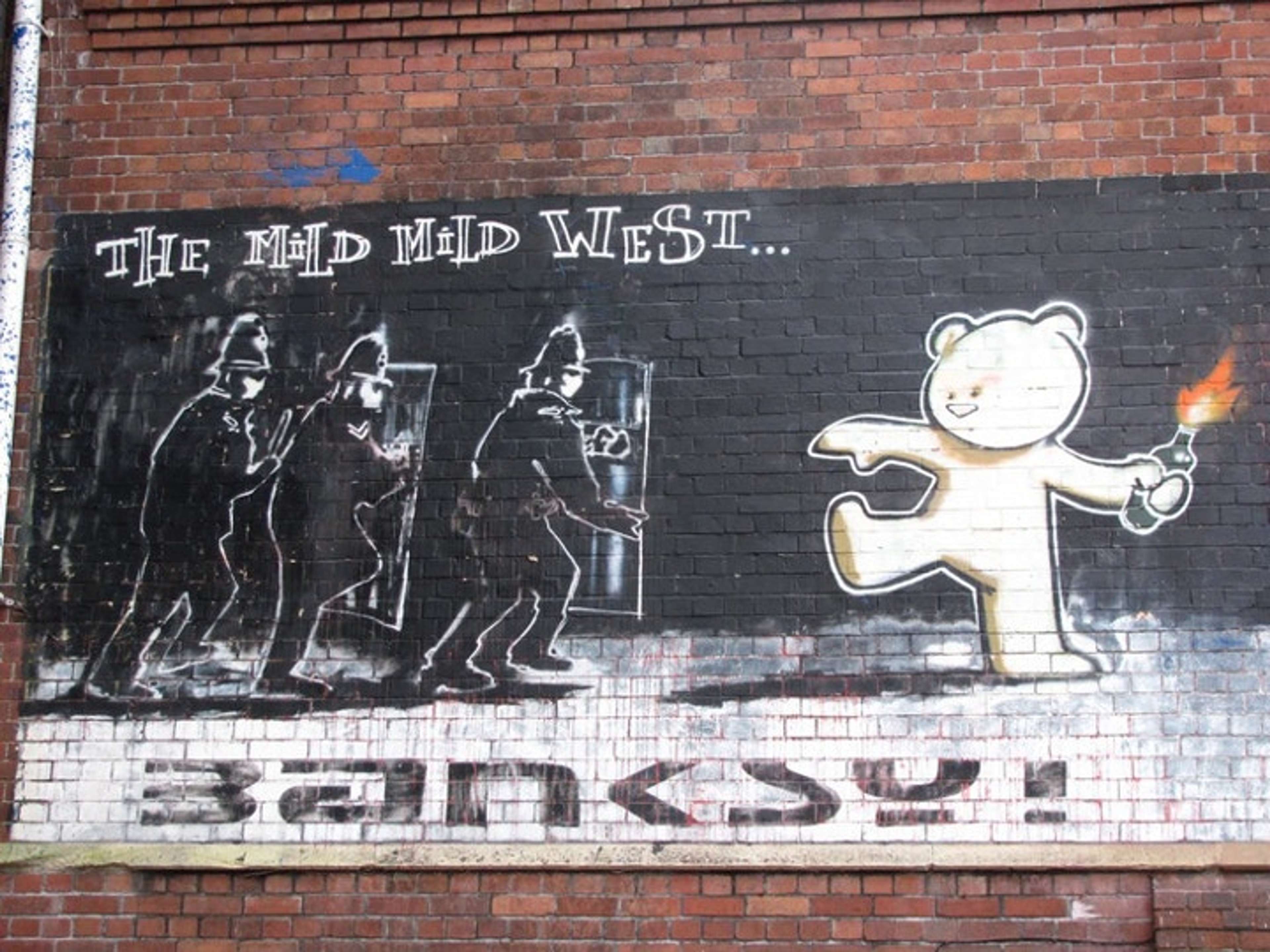 Une fresque en graffiti sur un mur de briques représente trois policiers anti-émeute avançant avec des boucliers, tandis qu'un grand ours en peluche blanc, de style dessin animé, marche vers eux tenant un cocktail Molotov allumé, sous le titre crayeux « THE MILD MILD WEST… ».