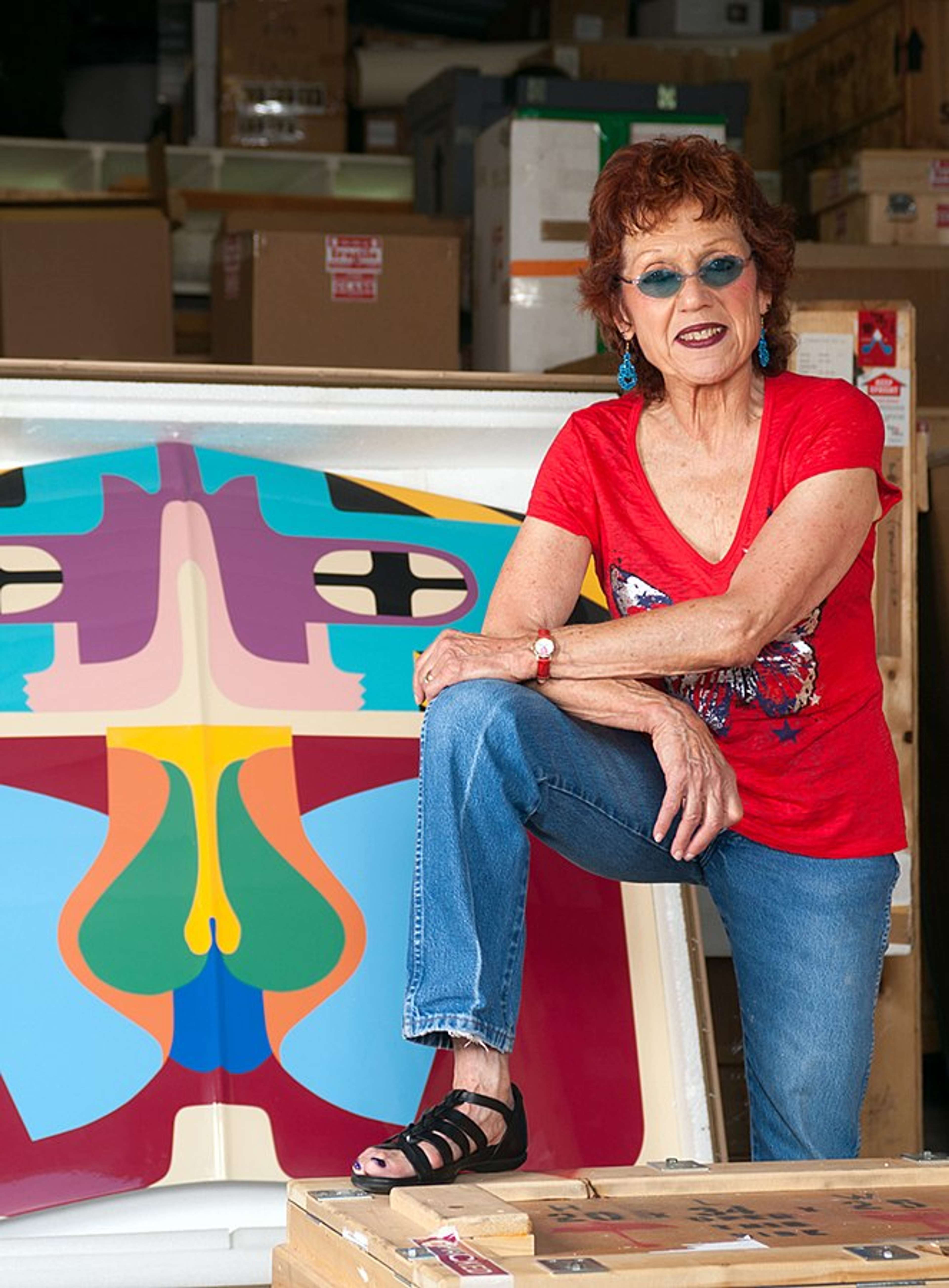 Une femme souriant à l'appareil photo, vêtue d'un haut rouge vif, entourée d'œuvres d'art