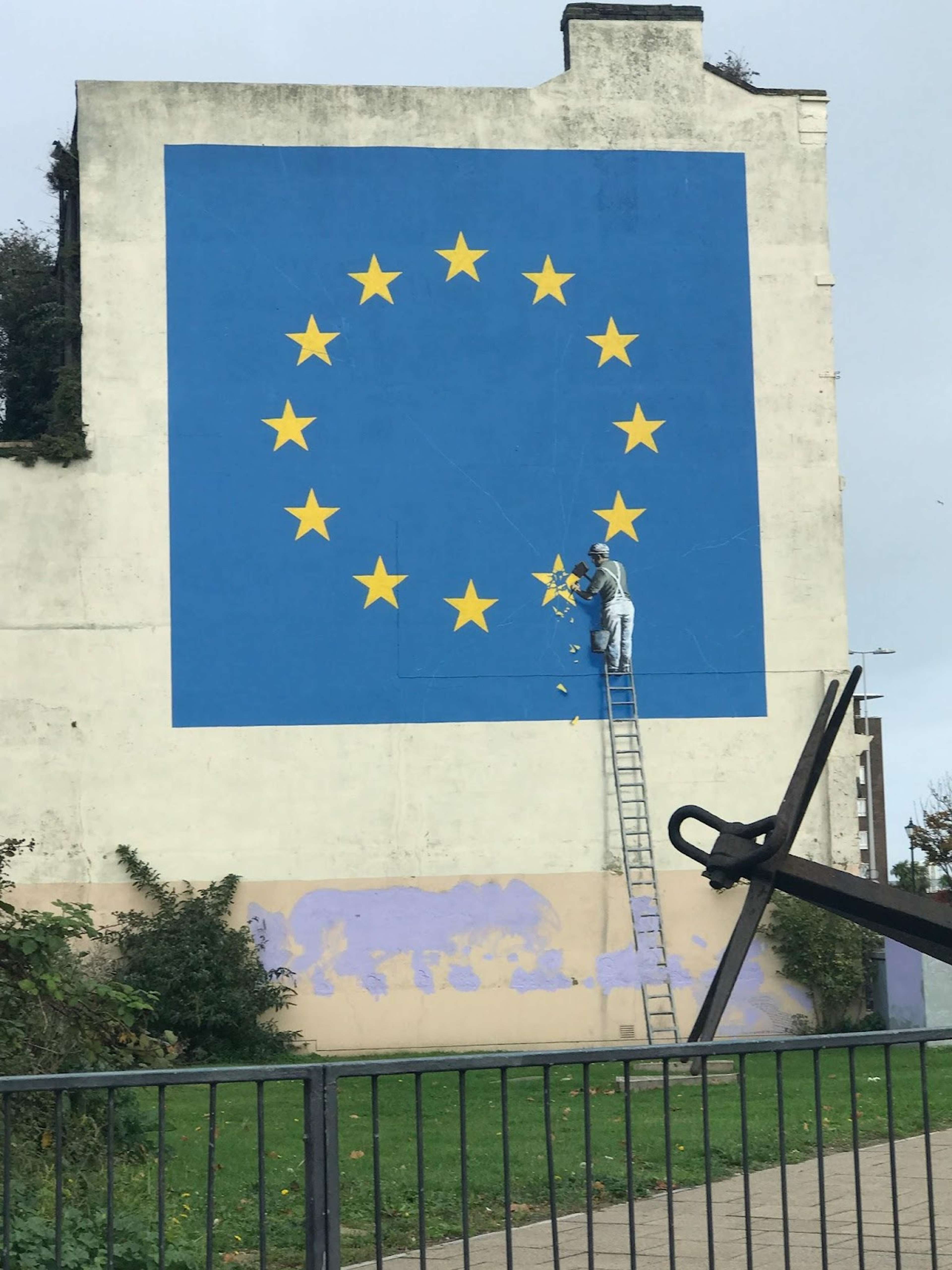 Une image de la fresque du Brexit de Banksy. Elle représente un grand drapeau de l'Union Européenne, en couleur, avec un homme monochrome sur une échelle en train d'en retirer une étoile.