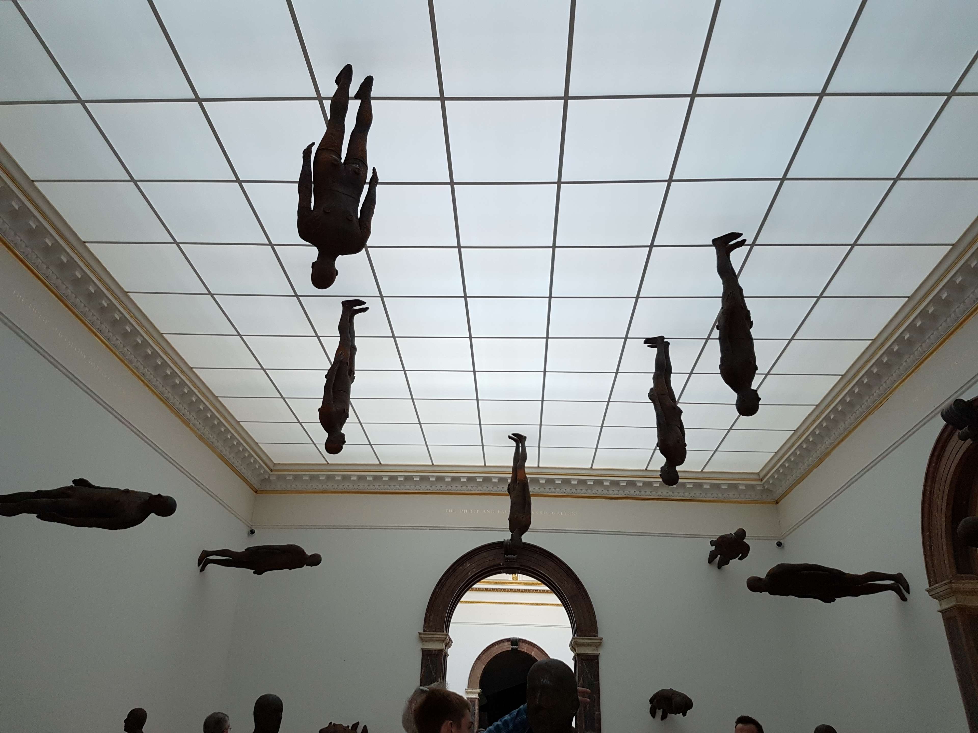 Une installation sculpturale d'Antony Gormley, composée de multiples figures en fonte grandeur nature suspendues à l'envers au plafond d'un espace de galerie classique. L'œuvre représente des formes humaines dans des orientations variées, leurs silhouettes rigides contrastant avec le plafond quadrillé de lumière et les moulures architecturales ornementées. L'installation explore la perception spatiale et la relation entre le Human Body et son environnement, exécutée dans le minimalisme figuratif caractéristique de Gormley avec une patine brun bronze discrète.