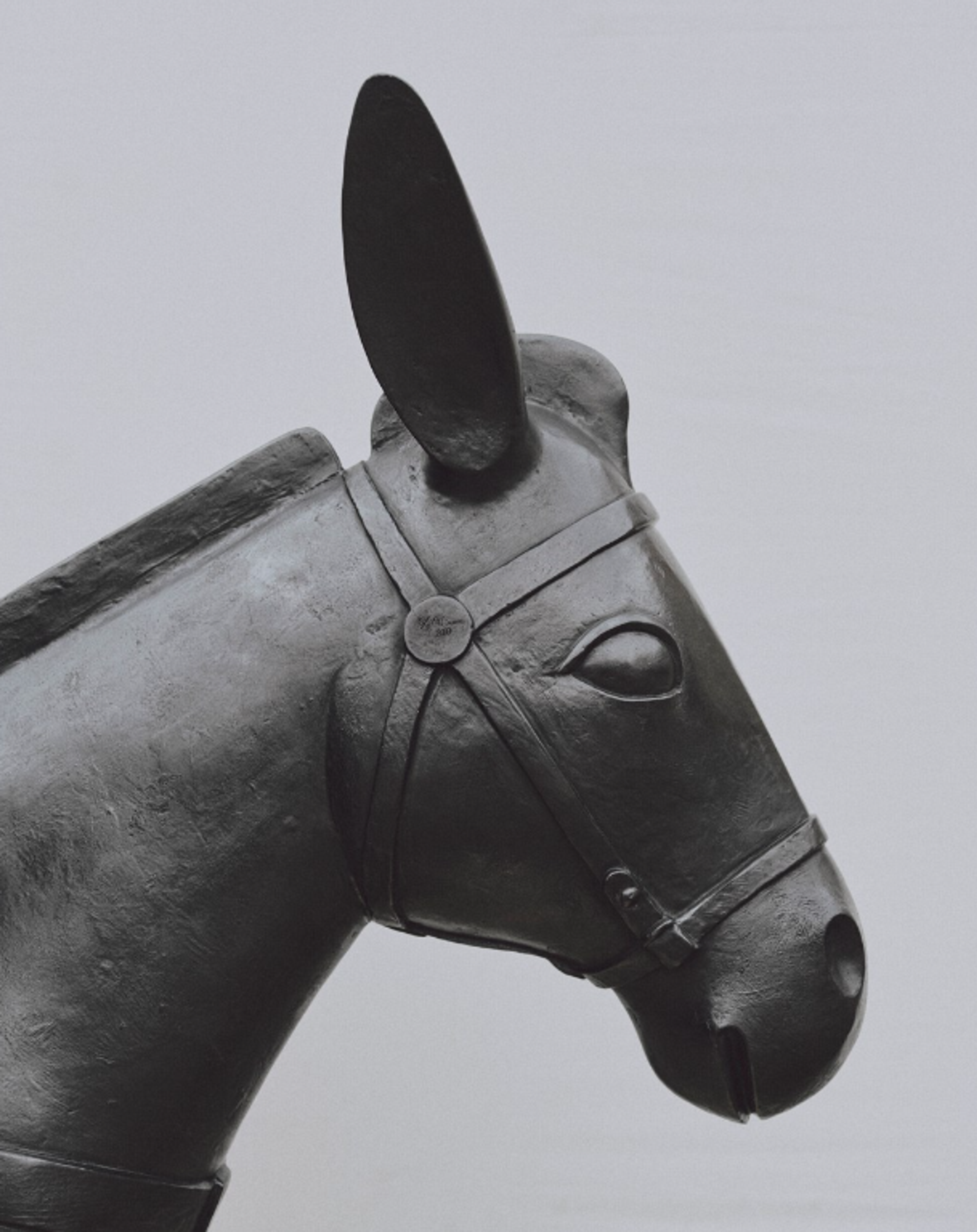 A bronze sculpture of a donkey carrying baskets of real white flowers on either side of his back. His form is simplified with smooth contours. The piece has a minimalist aesthetic with subtle facial features and alert ears. 
