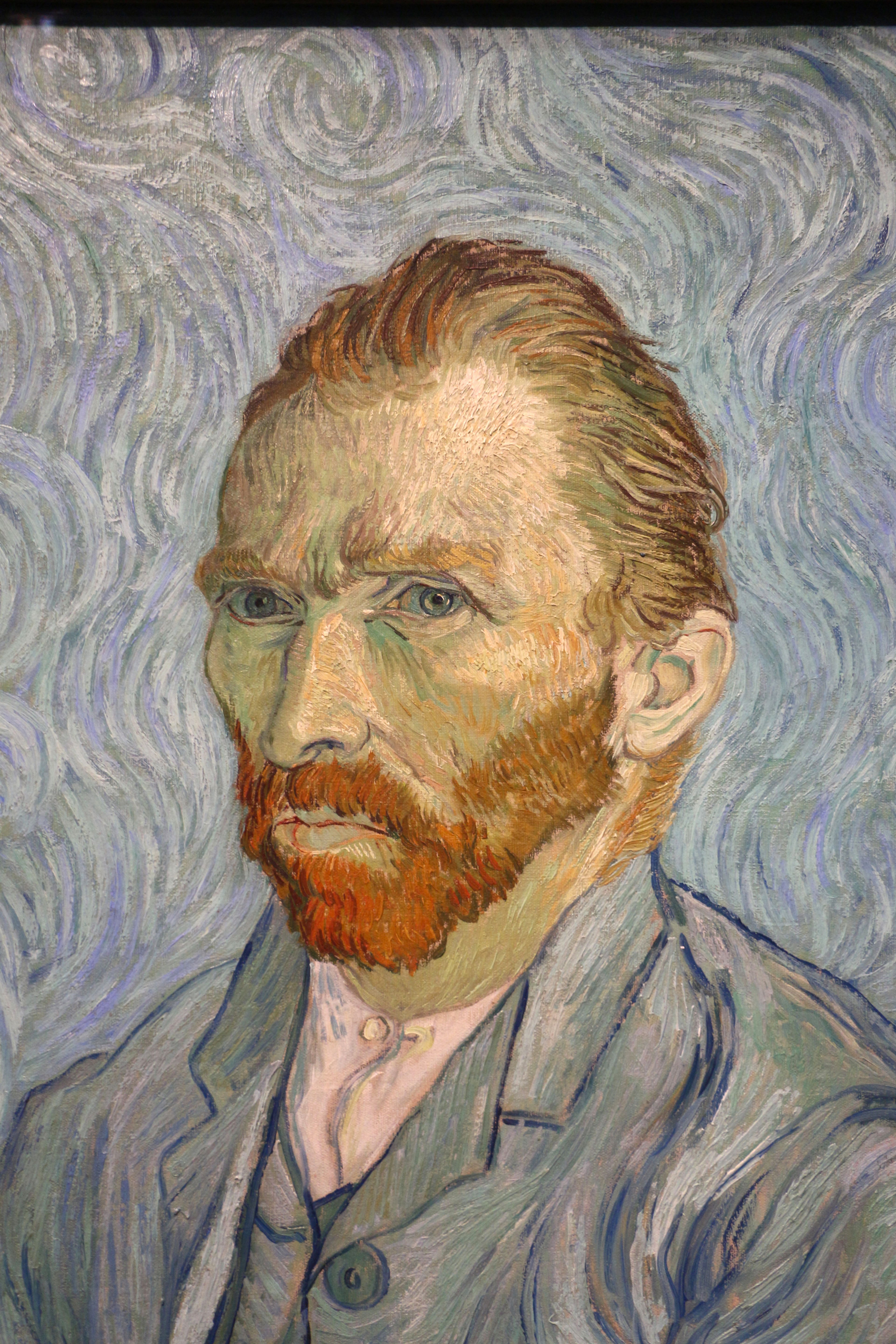 Un autoportrait de Vincent Van Gogh, avec sa chevelure et sa barbe rousses caractéristiques. Il porte un costume bleu sur un fond bleu et regarde directement l'observateur.