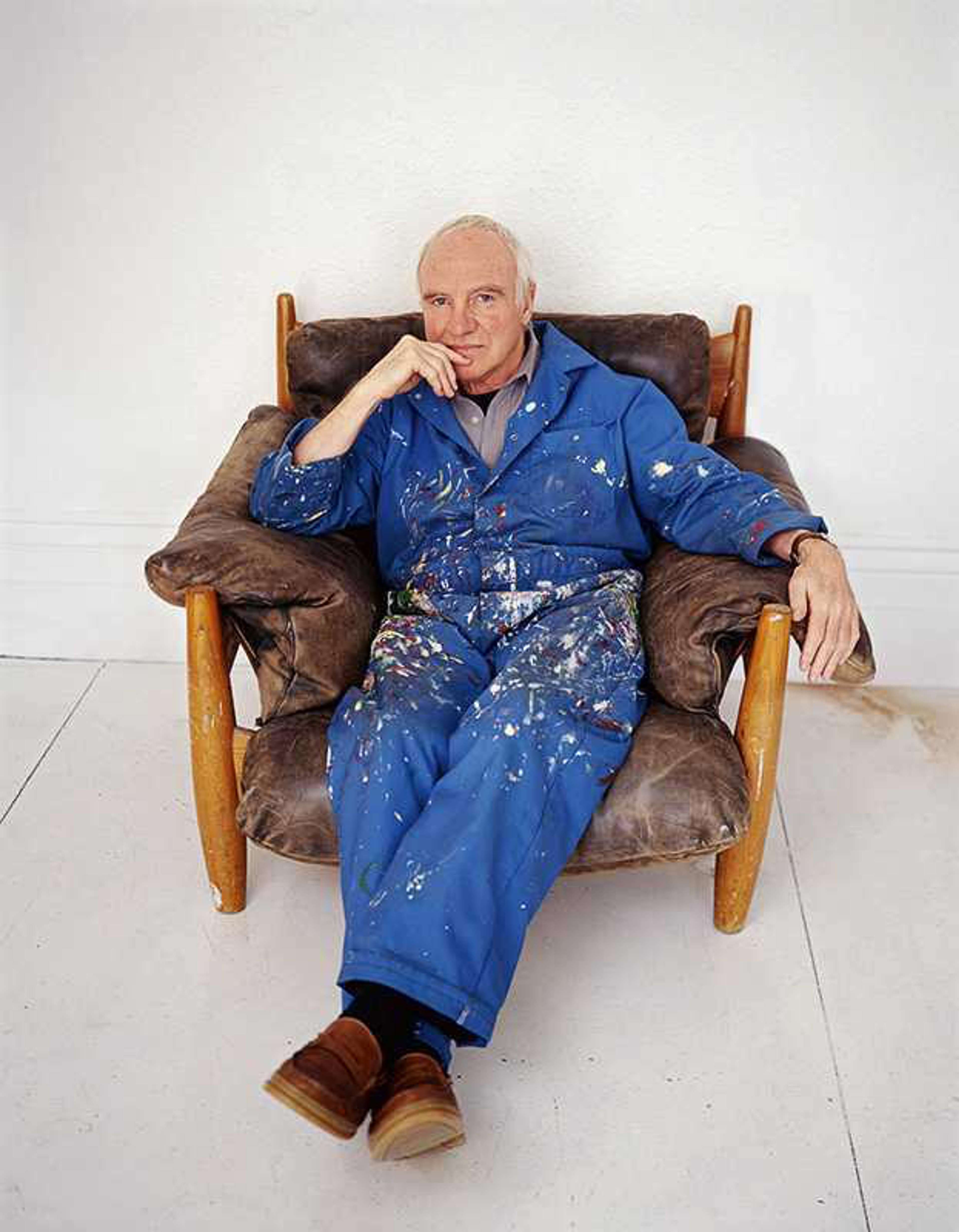 Photographie de l'artiste Patrick Caulfield vêtu d'une salopette éclaboussée de peinture, assis dans un fauteuil en cuir sur fond blanc.