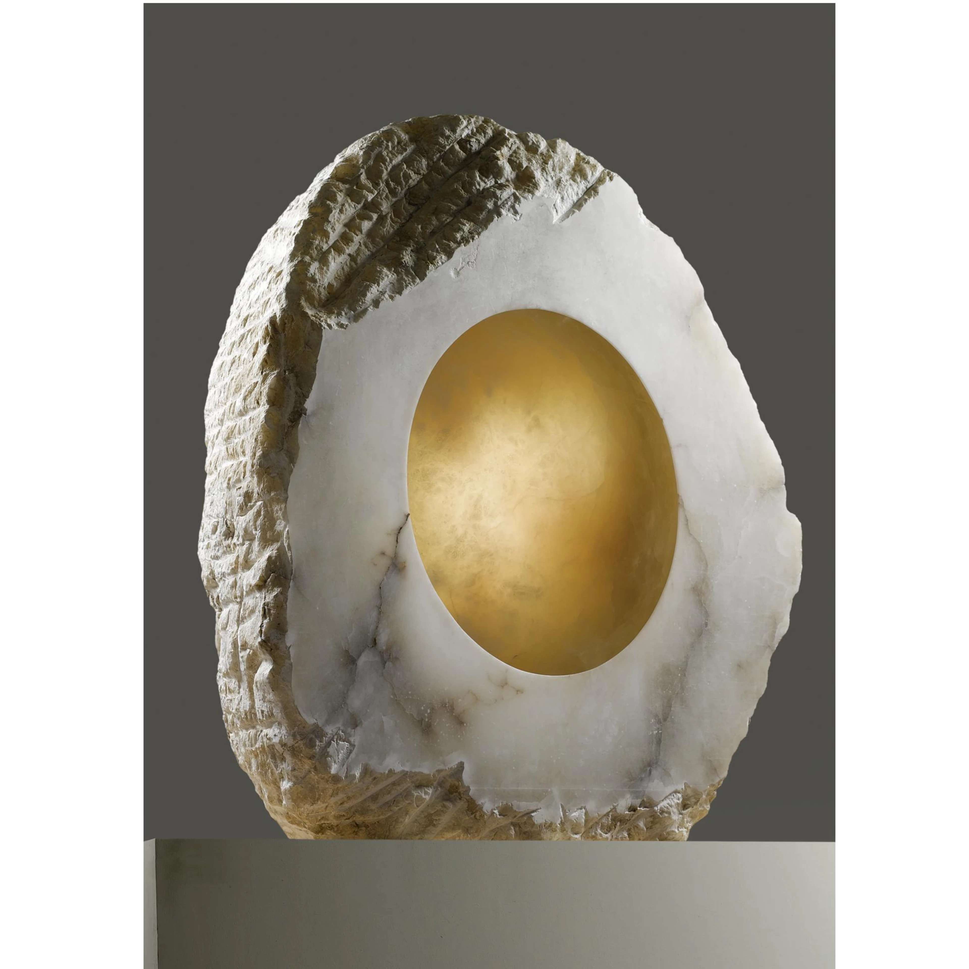 A roughly-hewn alabaster rock with jagged edges and one perfectly smooth vertical face. Out of the smooth face, a half-sphere has been carved, the insides of which are mottled gold. The alabaster rock has thick dark grey veins running through its translucent surface.