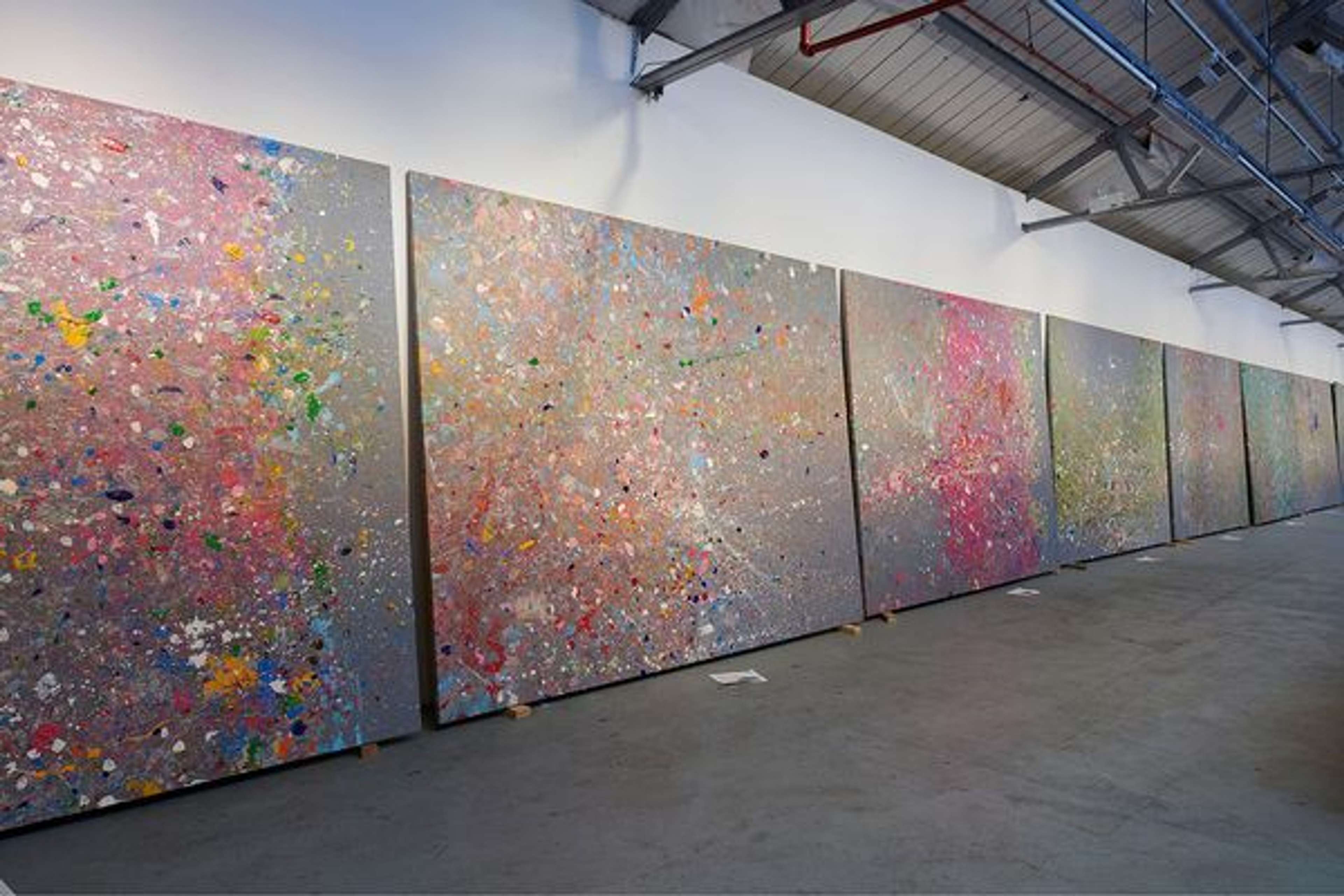 Une photographie de l'atelier de Damien Hirst, montrant plusieurs toiles de sa série « Coast Paintings ». Il s'agit d'une série d'éclaboussures de peinture colorées sur un fond gris.