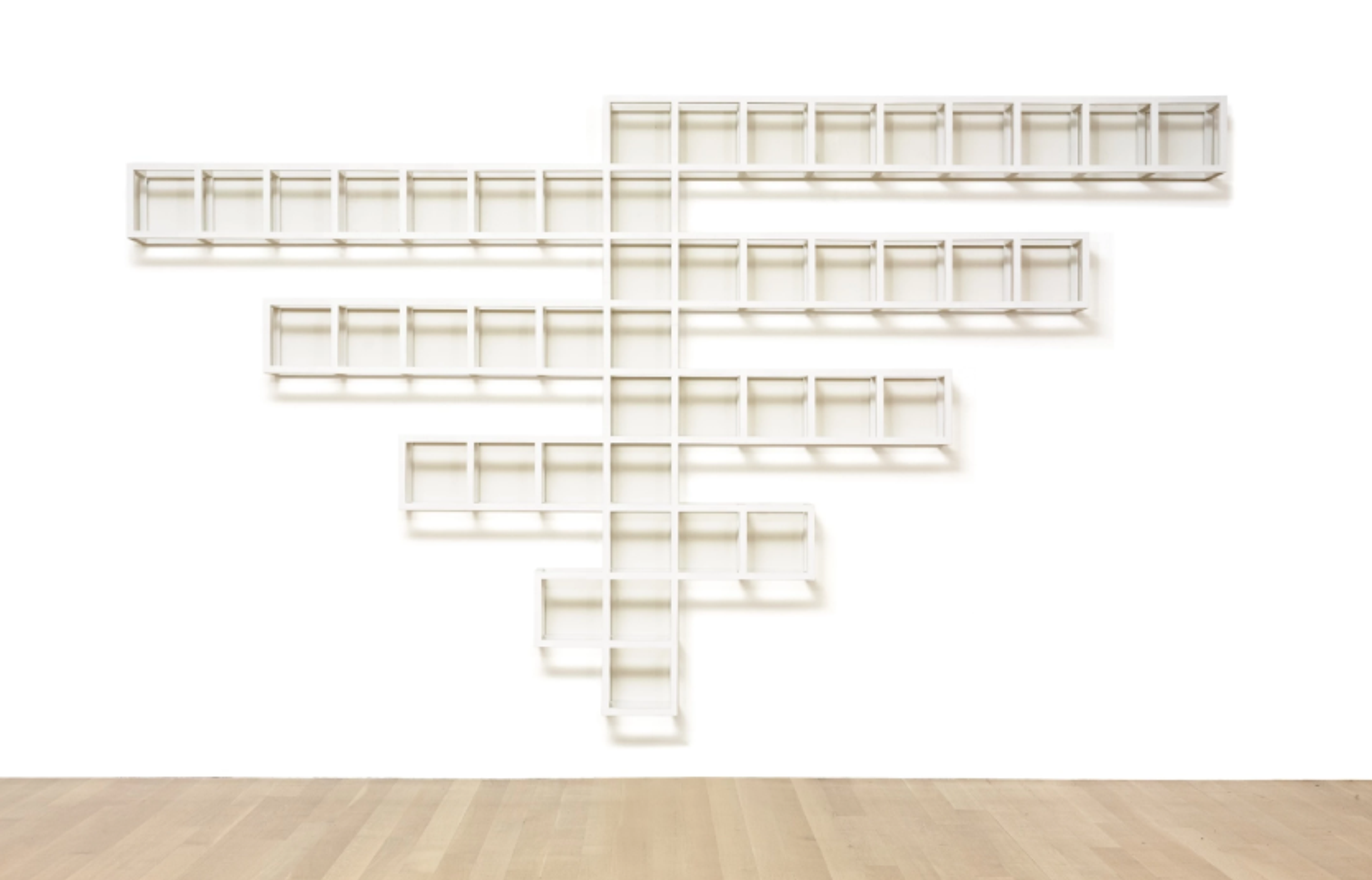 A geometric arrangement of open white cubes set against a white wall. The arrangement is only ever one cube deep. It has a central vertical line of cubes, with alternating straight branches of cubes which get longer as they get higher. The top branch is nine cubes long and sits on the right.