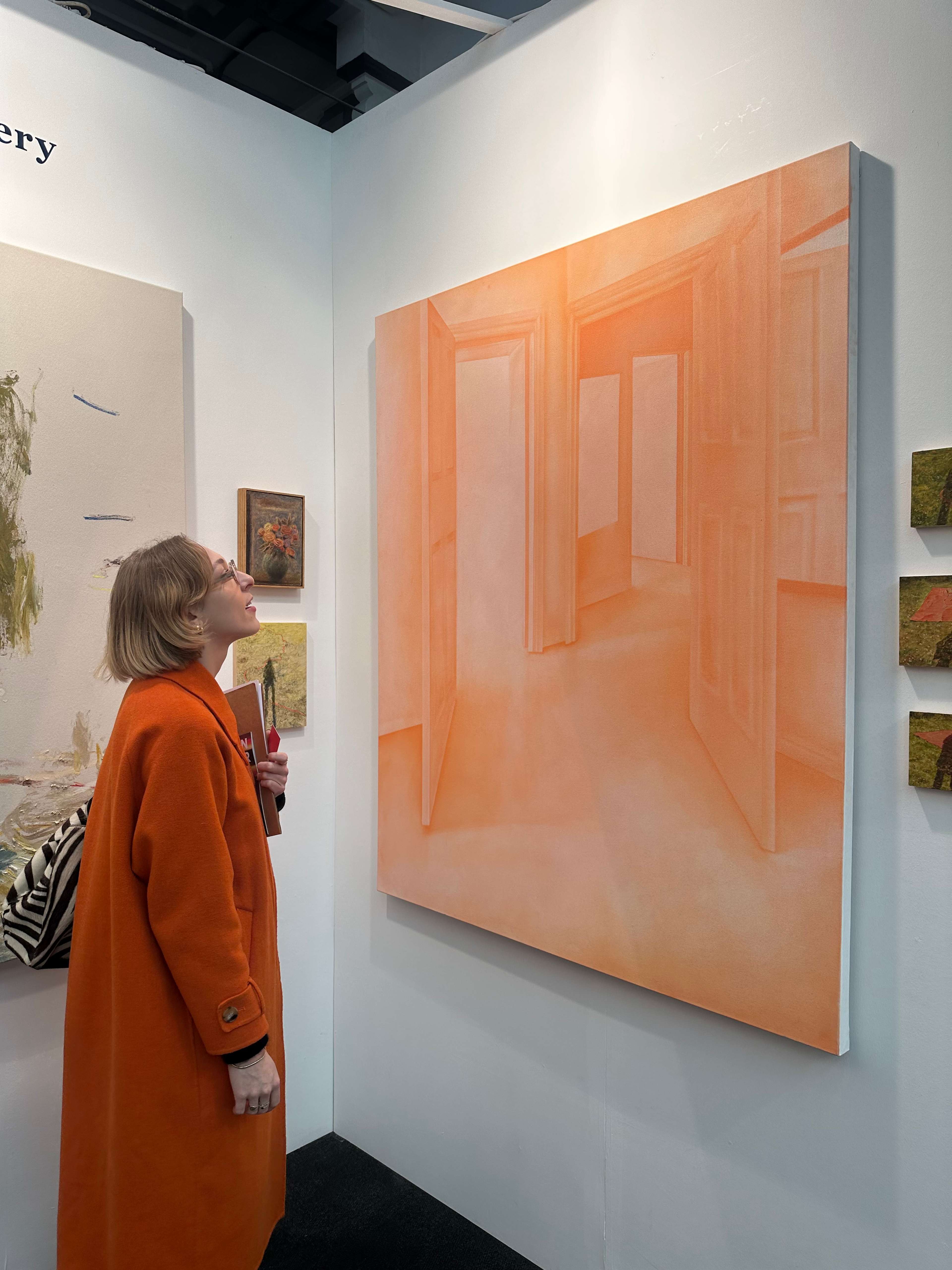 Une jeune femme vêtue d'un long manteau orange regarde vers le haut une grande toile accrochée au mur