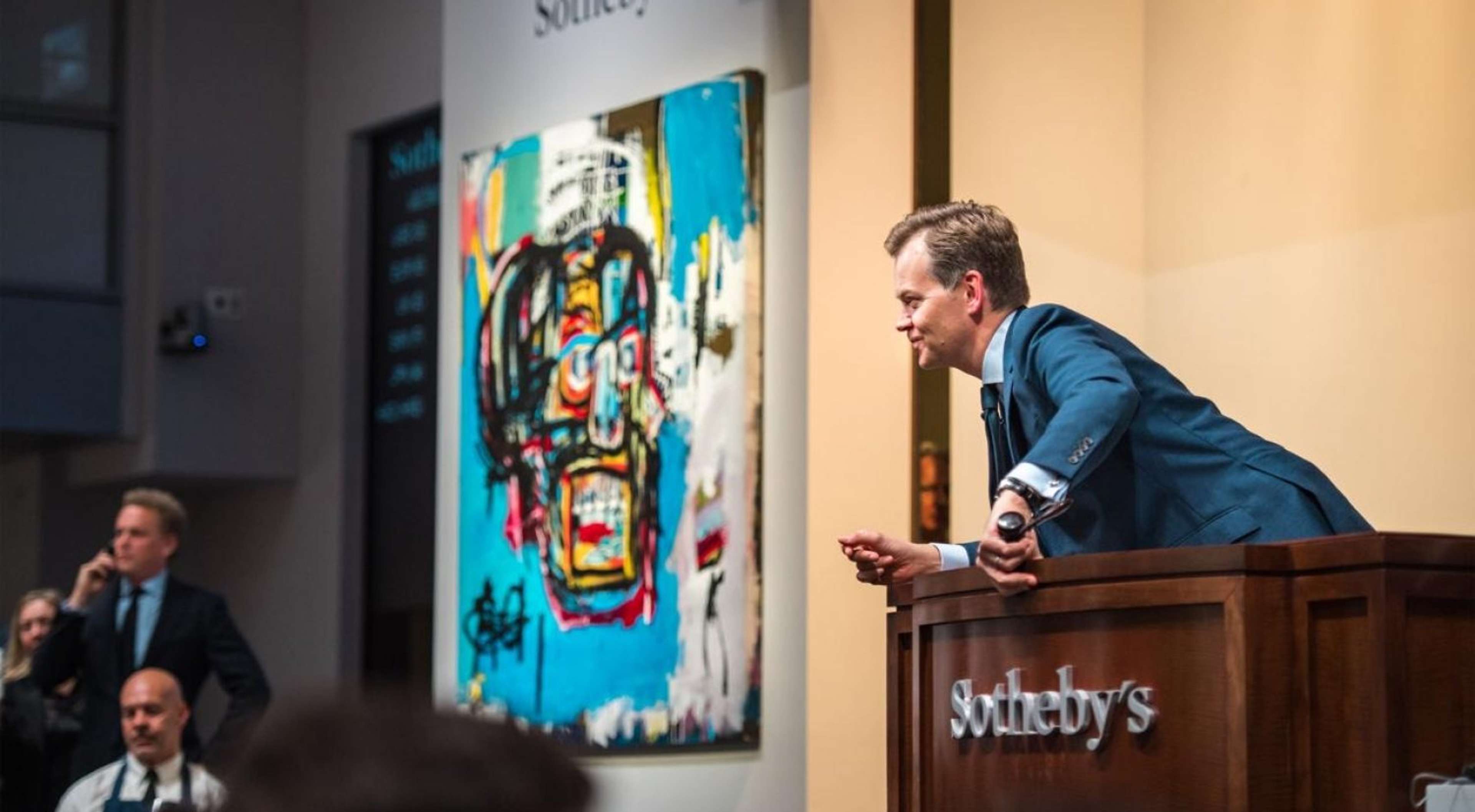 Une image montrant un commissaire-priseur en interaction avec la foule lors d'une vente aux enchères Sotheby's. En arrière-plan, on distingue l'œuvre "Untitled" de Basquiat datant de 1982.
