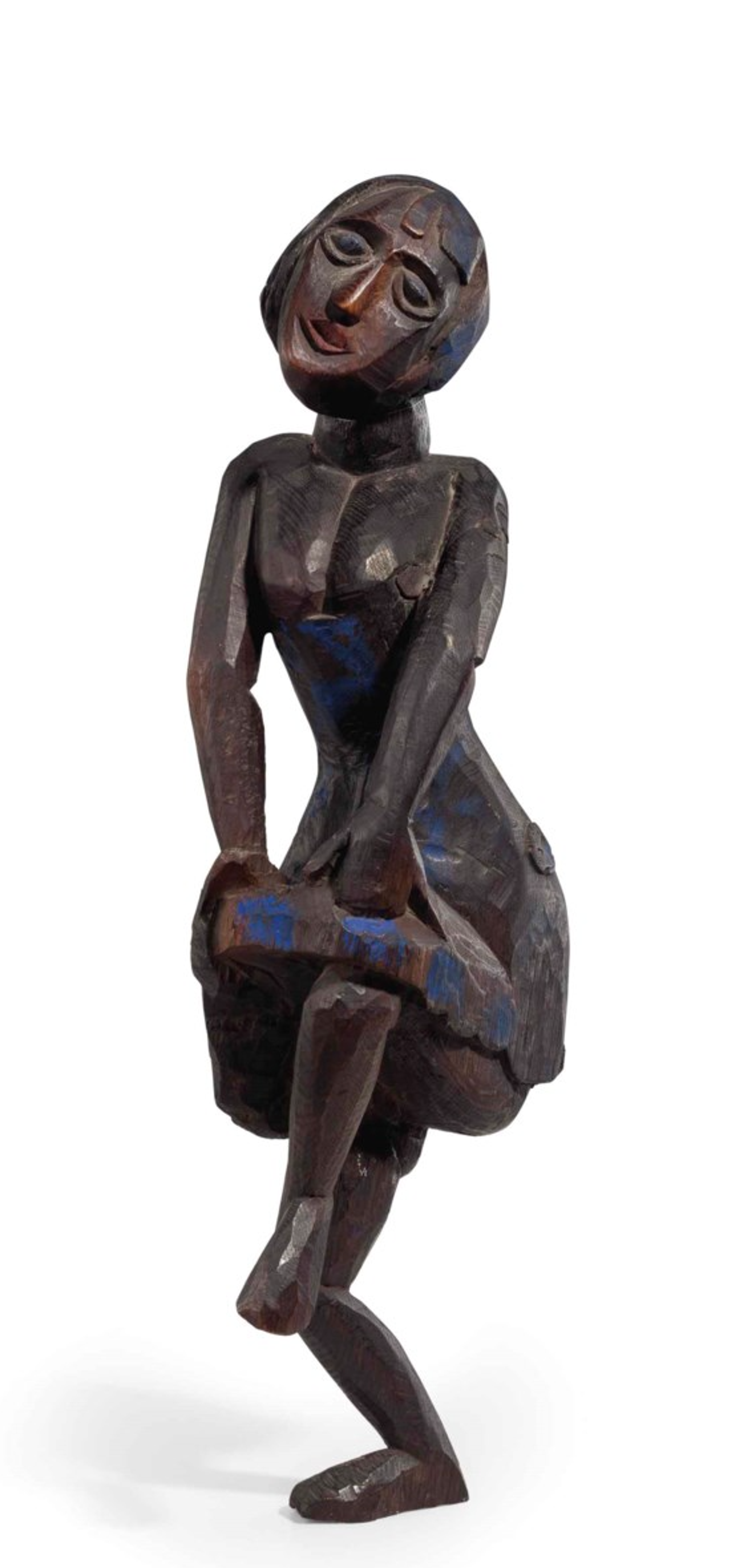 An oak sculpture standing approximately 67 cm tall, depicting a female dancer holding her skirt with two hands above her one raised leg. Both legs are bent at the knee. Her head is tilted up and to the side. The figure has been carved with elongated proportions and simplified features, including a mask-like face. The surface of the oak is painted dark and glossy. 