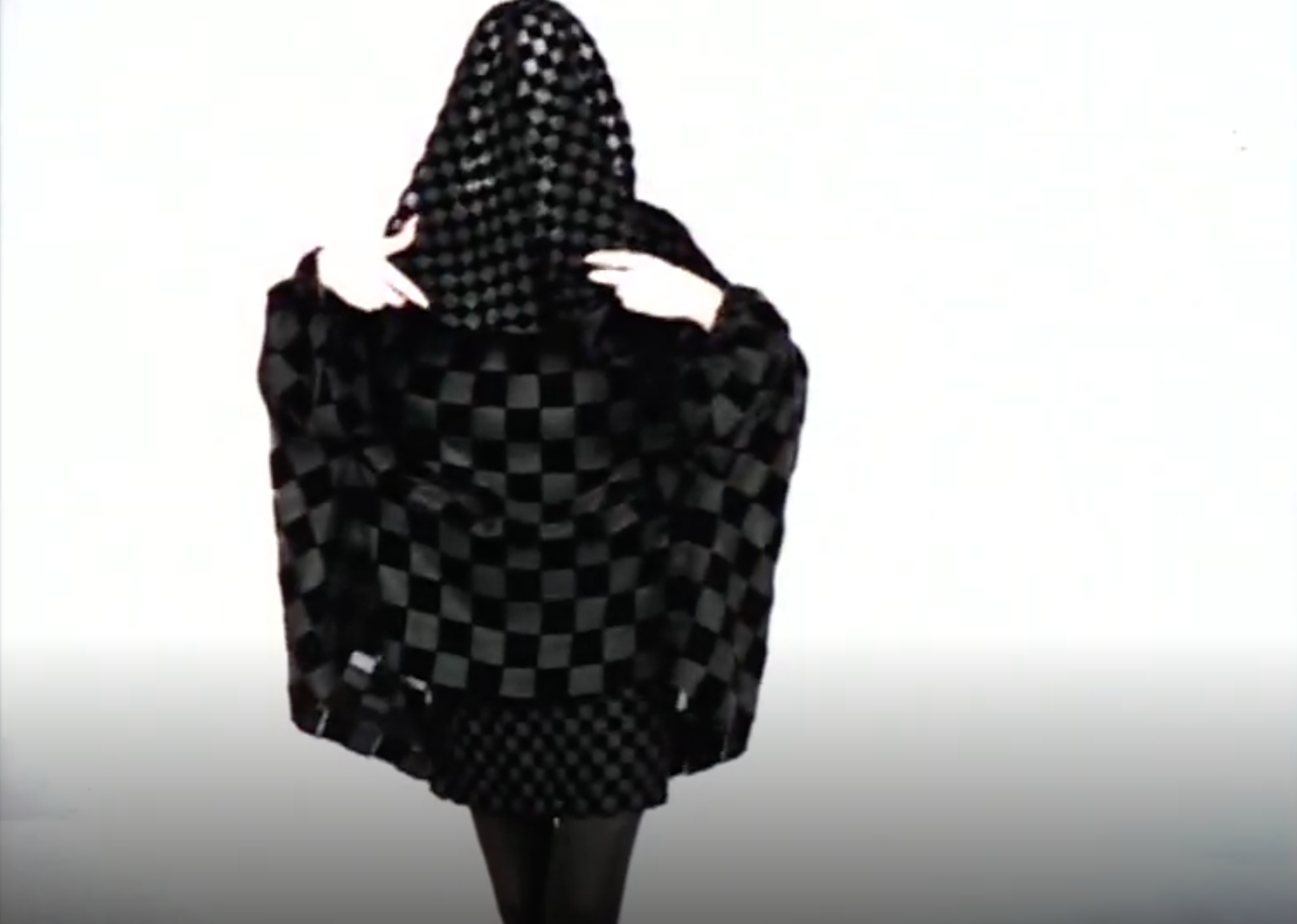 Une capture d'écran d'une femme voilée vêtue de noir sur fond blanc. Sa tenue entière est ornée de motifs de carrés noirs de différentes tailles.