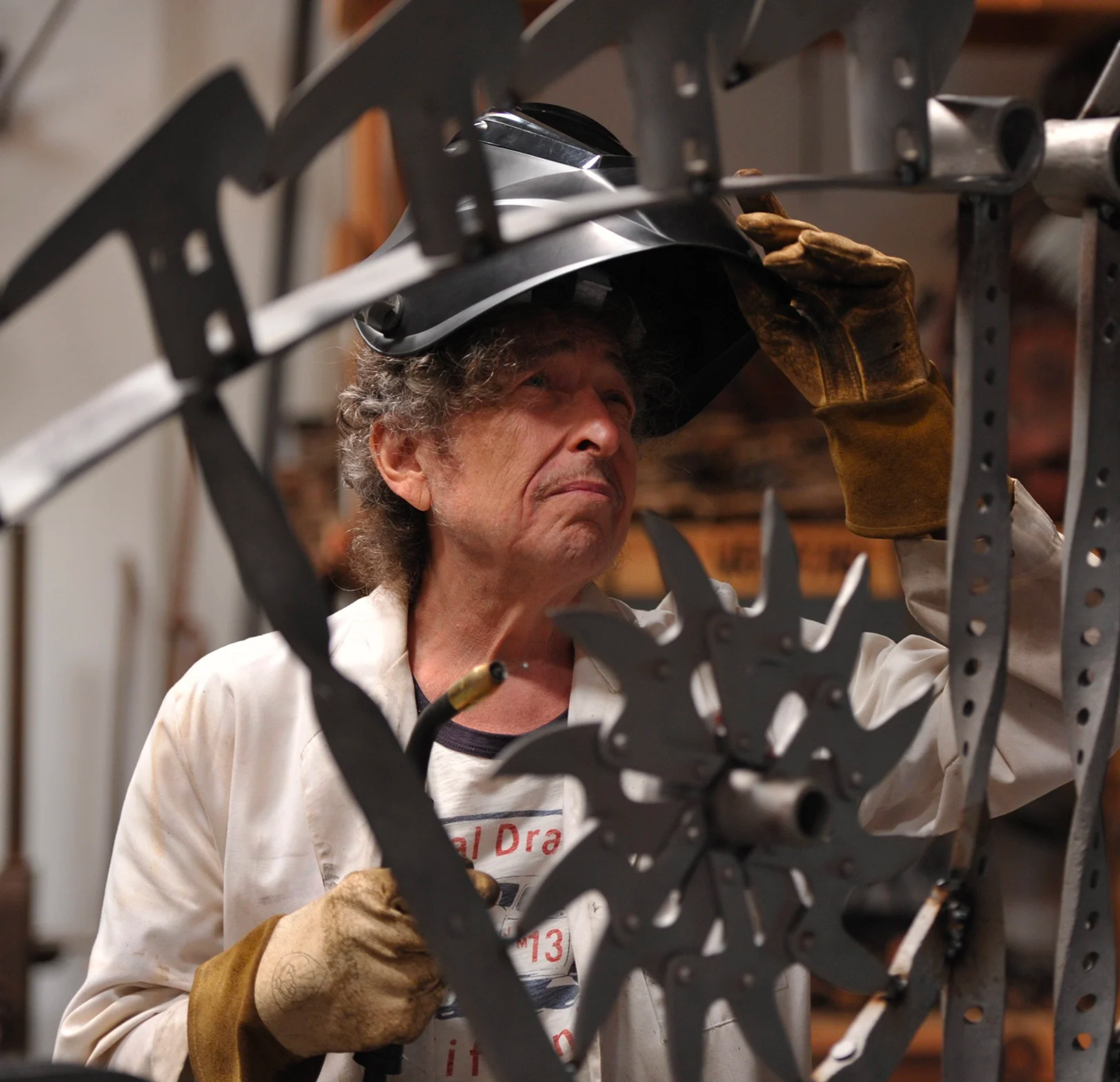 Photographie de Bob Dylan portant des gants jaunes et un casque de chantier, travaillant dans son atelier sur une sculpture en métal.