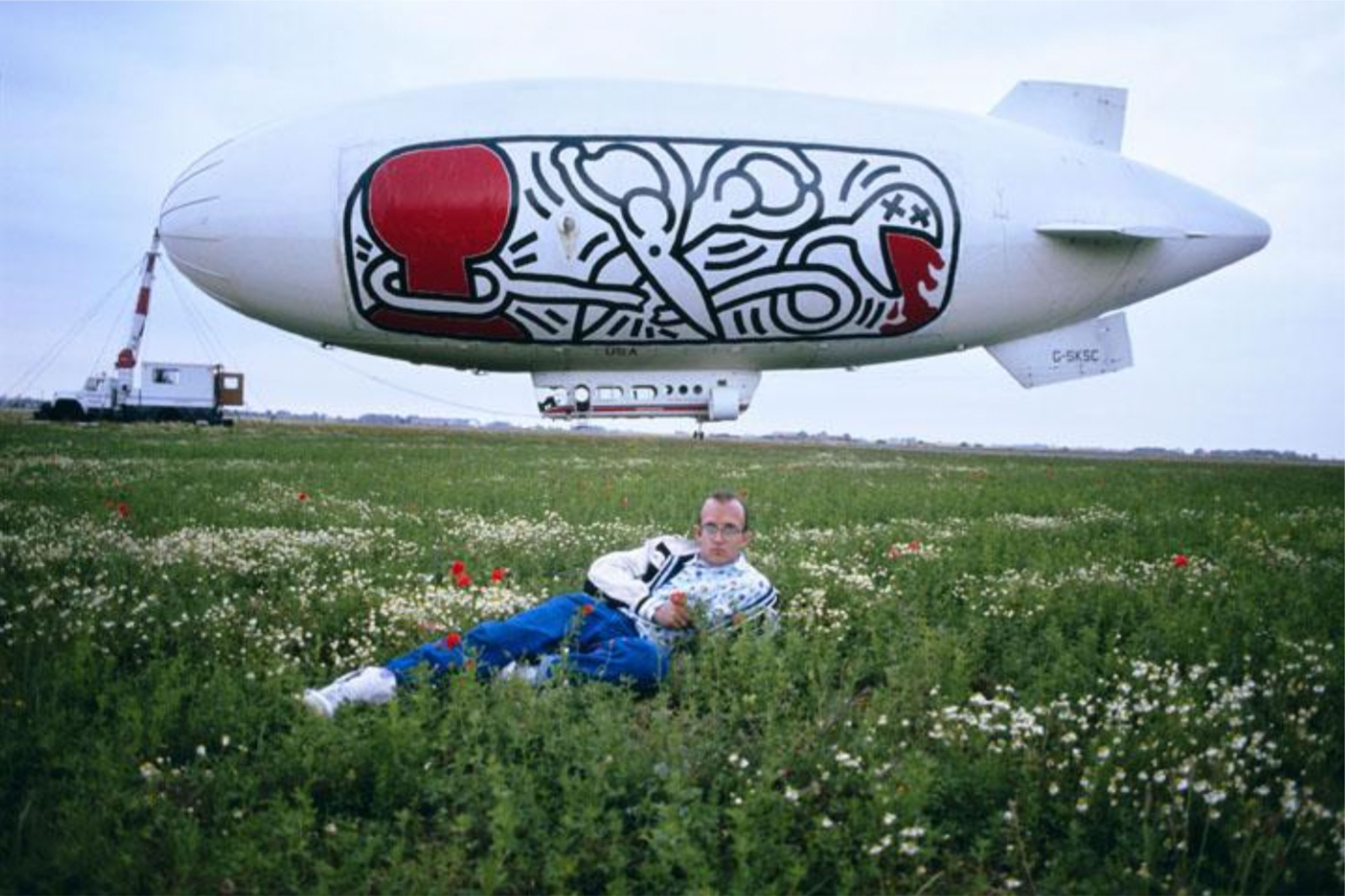 Keith Haring allongé dans un champ d'herbe, avec un dirigeable arborant ses œuvres derrière lui en France.