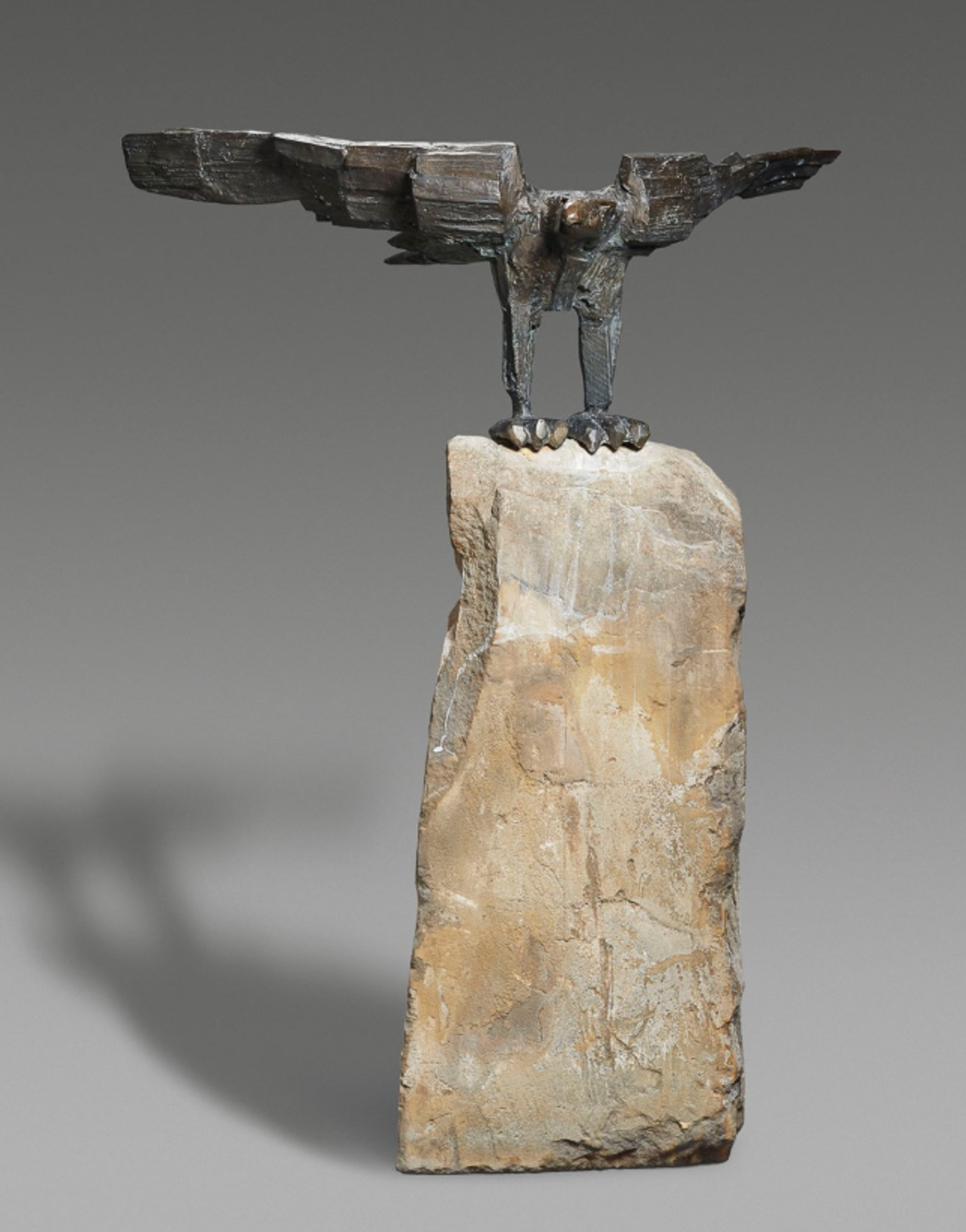 A rugged piece of mottled brown stone, roughly cut into a vertical cuboid. On top, sits a bronze lectern shaped like an eagle. The eagle is asymmetrical, with uneven legs, a rough surface, and block-like wings.