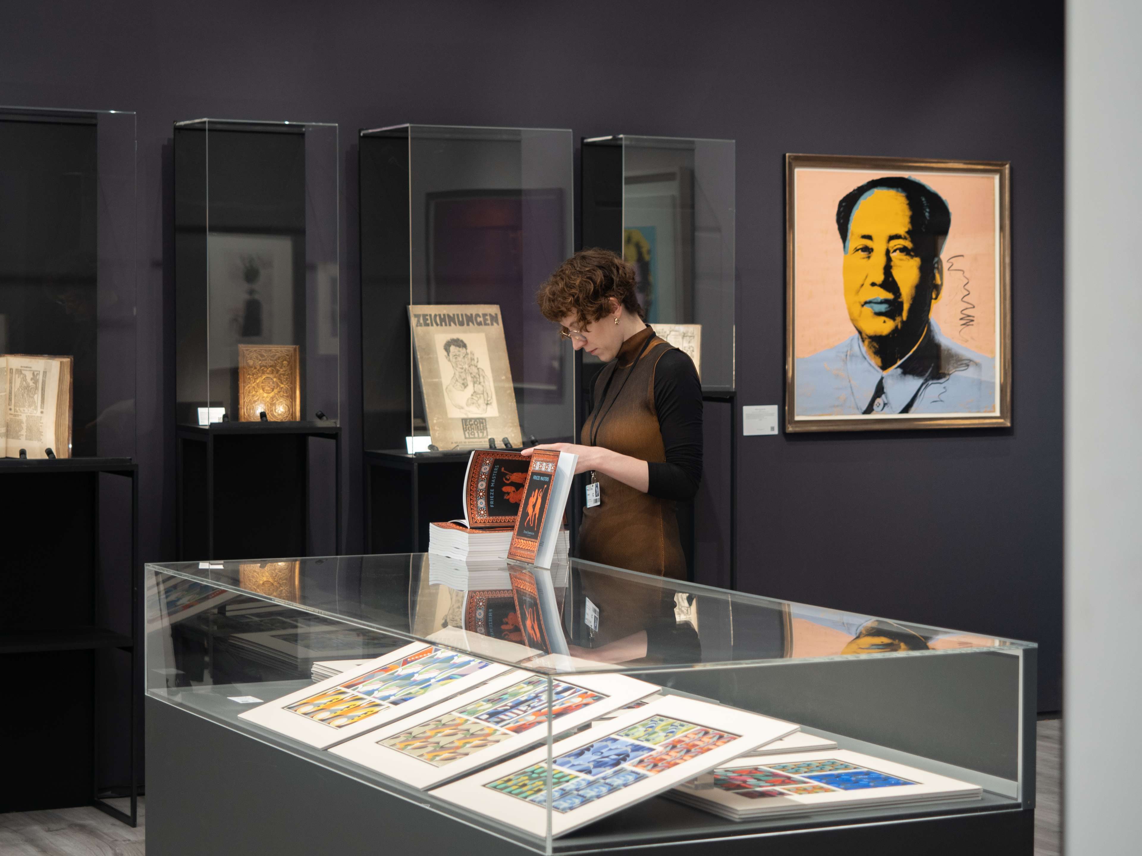 Image d'une femme feuilletant un magazine à côté d'un aquarium en verre blanc dans un stand de Frieze Masters 2023, avec l'œuvre d'Andy Warhol intitulée 'Mao' accrochée au mur.