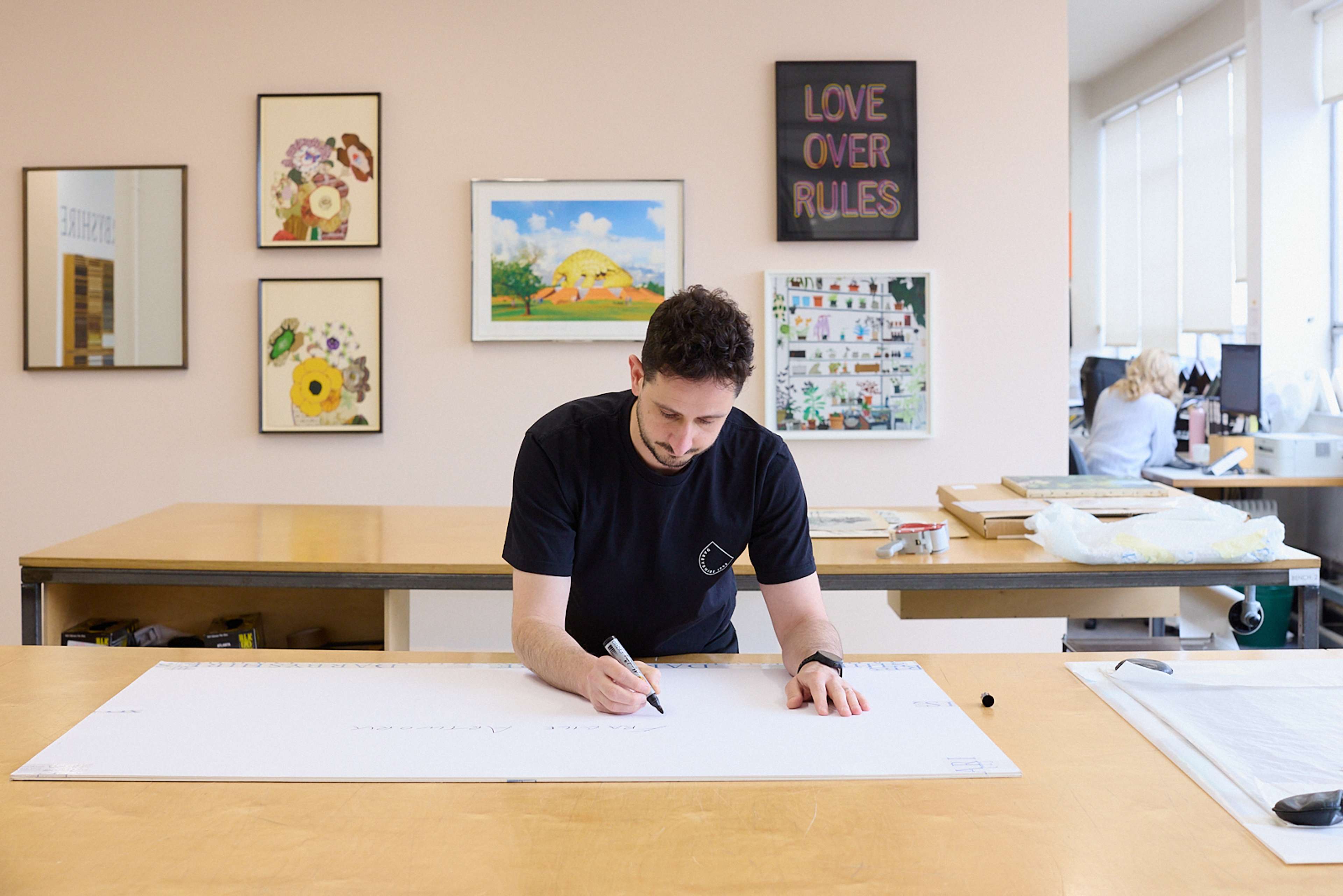 Consultant Darbyshire écrivant sur un grand panneau de montage à une table de travail dans "In The Studio", avec des œuvres encadrées visibles sur "The Wall" derrière.
