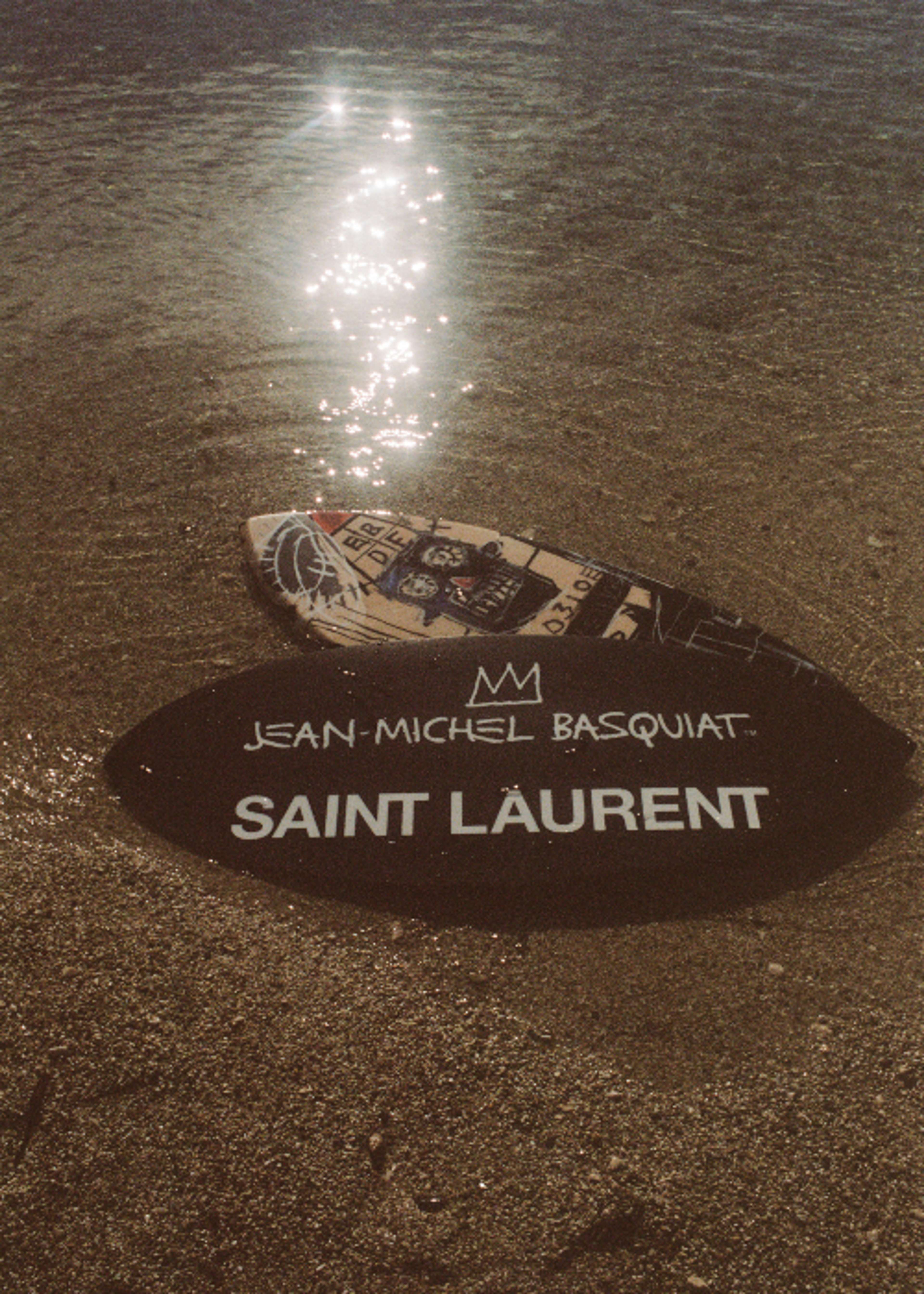 Une image de deux skimboards posés sur le sable, chacun montrant une face de l'objet. Le dessus présente une œuvre de Basquiat, tandis que le dessous de la planche porte le nom de l'artiste surmonté d'une couronne ainsi que le logo Saint Laurent.