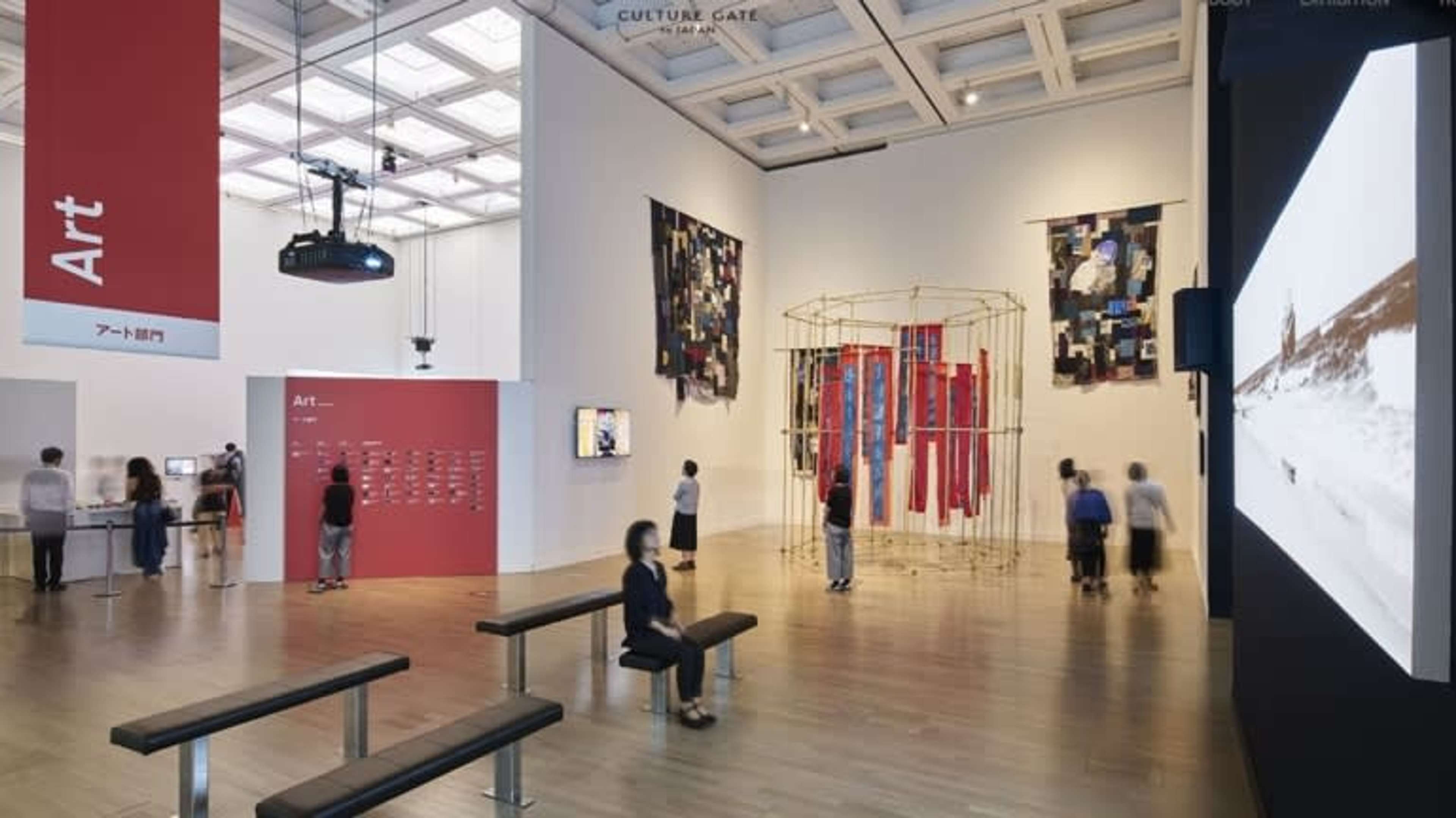 Espace de galerie avec une femme assise regardant une installation vidéo et des œuvres d'art à l'arrière-plan et dans les environs