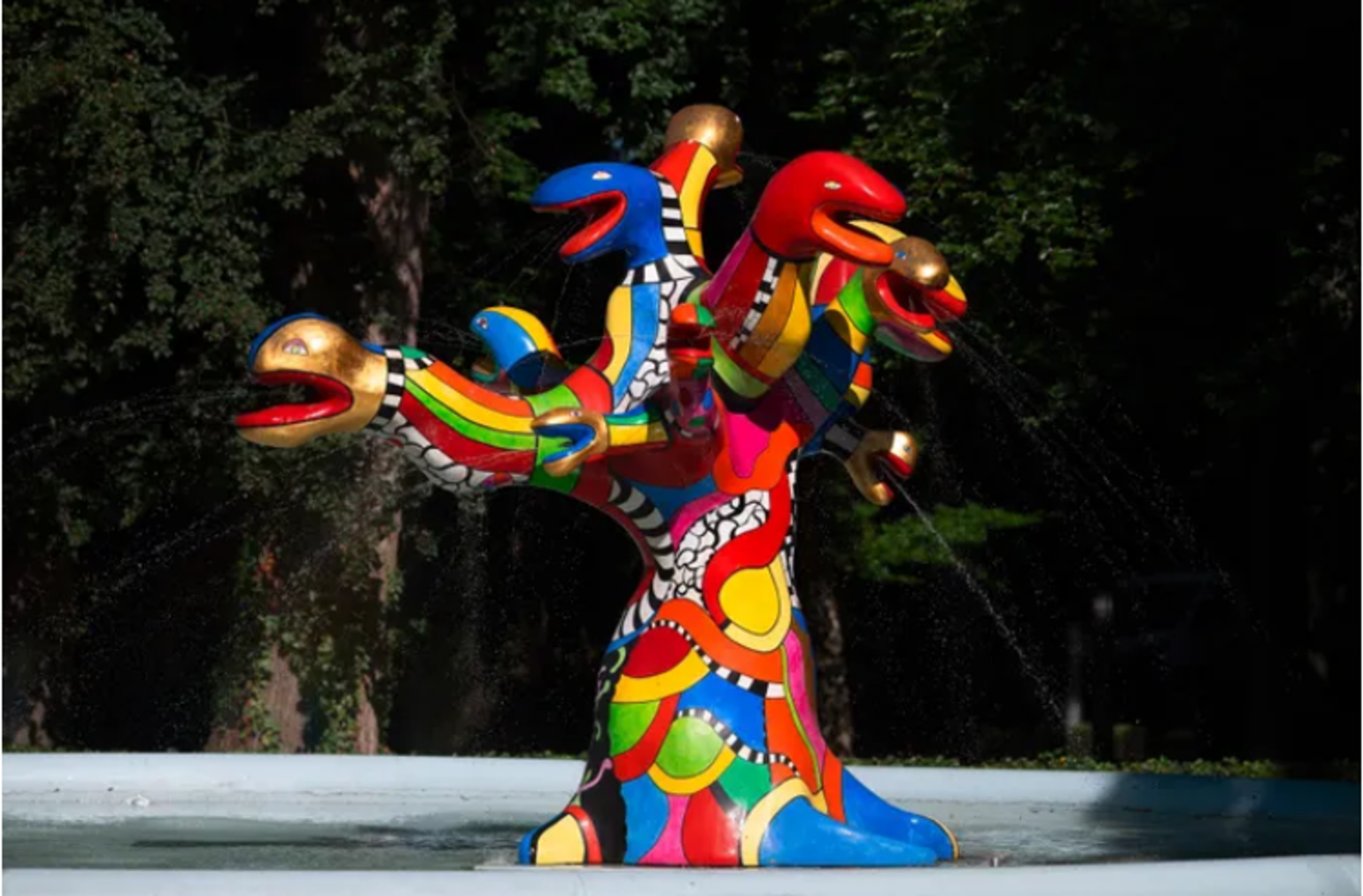 A bold outdoor sculpture featuring a tree-like form with multiple colourful, serpentine heads or branches. The piece combines bright primary colors with striped patterns and features dynamic, curving forms.
