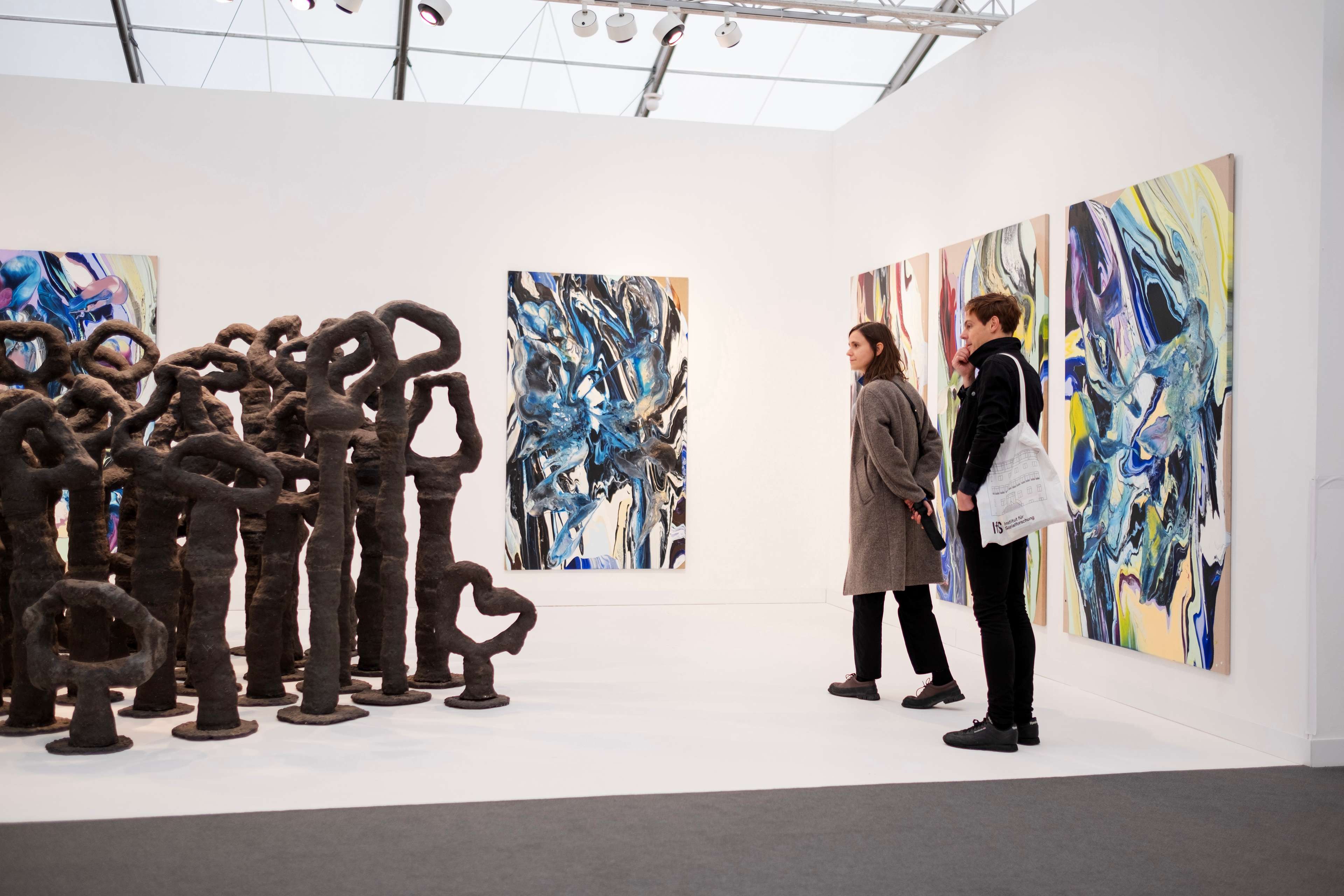 Image de deux personnes debout dans un stand de galerie lors de la foire Frieze London 2023, observant une collection d'installations sculpturales avec de grandes peintures murales ornant les murs du stand.