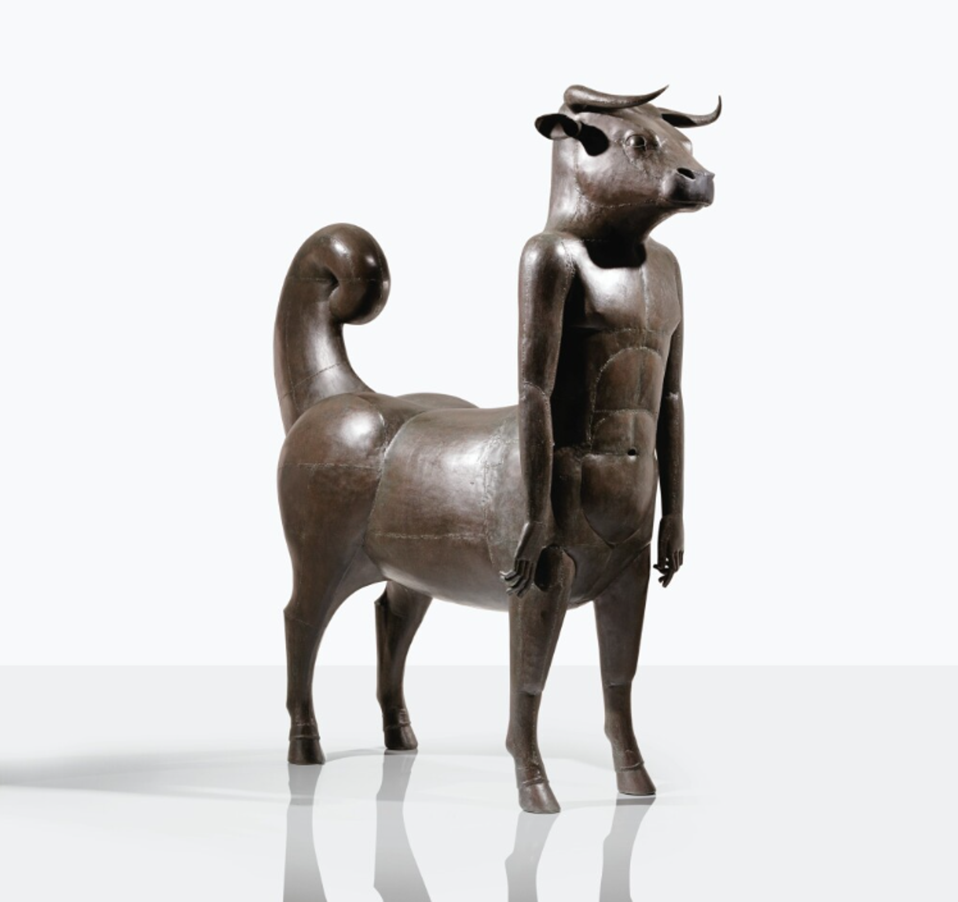A bronze sculpture depicting the mythological Minotaur. The figure stands upright on four legs with a stylised, elongated form and smooth texture. It has a human torso and a bull’s head. Its arms are very long, and the muscles of its abdomen are established by the connecting joins of sheet metal. It has a thick, curling tail.