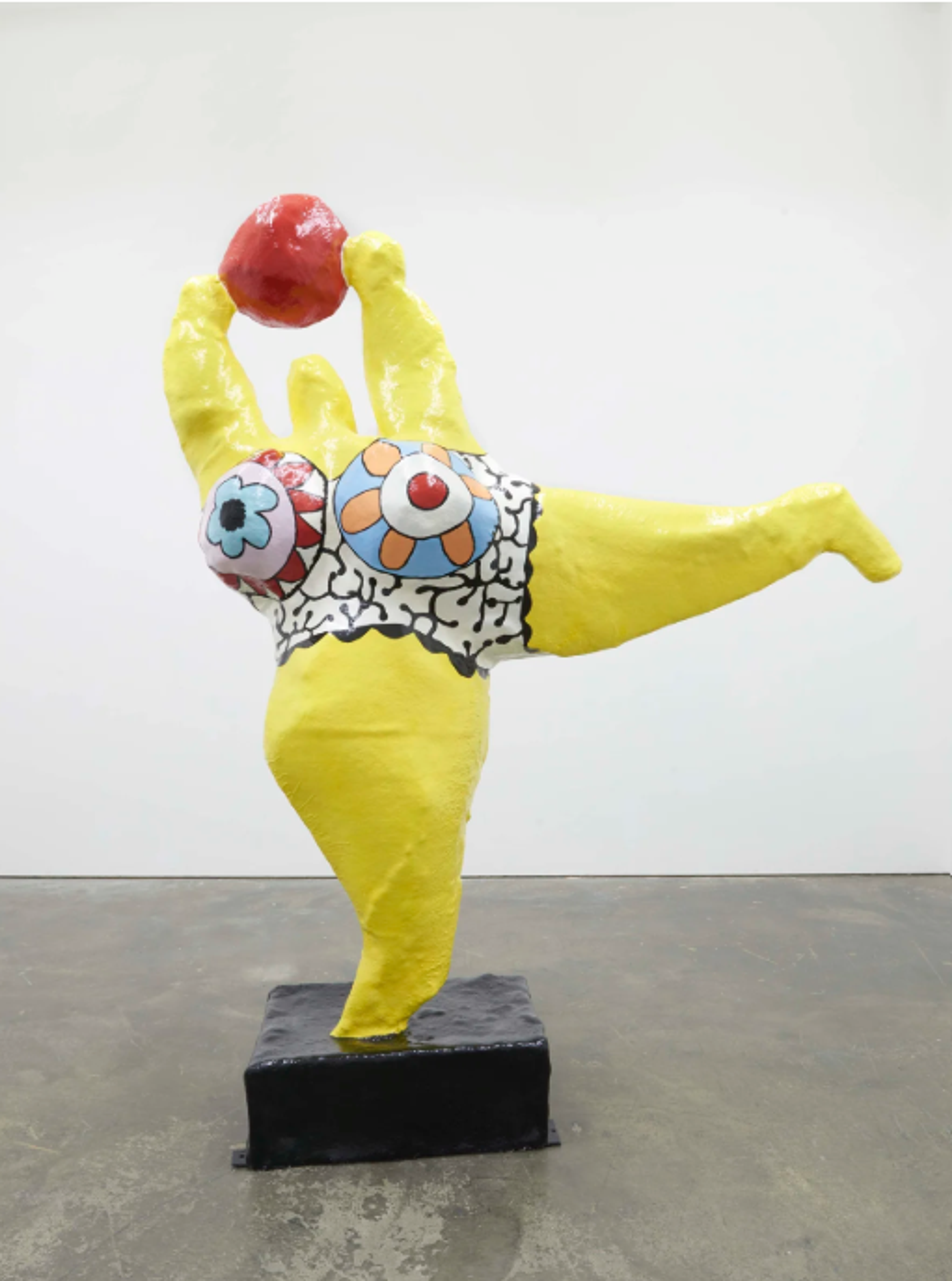 A joyful yellow sculptural female figure balances gracefully on one leg while holding a red ball aloft. The figure wears a patterned costume featuring circular designs in blue and white, embodying movement and playfulness in its dynamic pose.