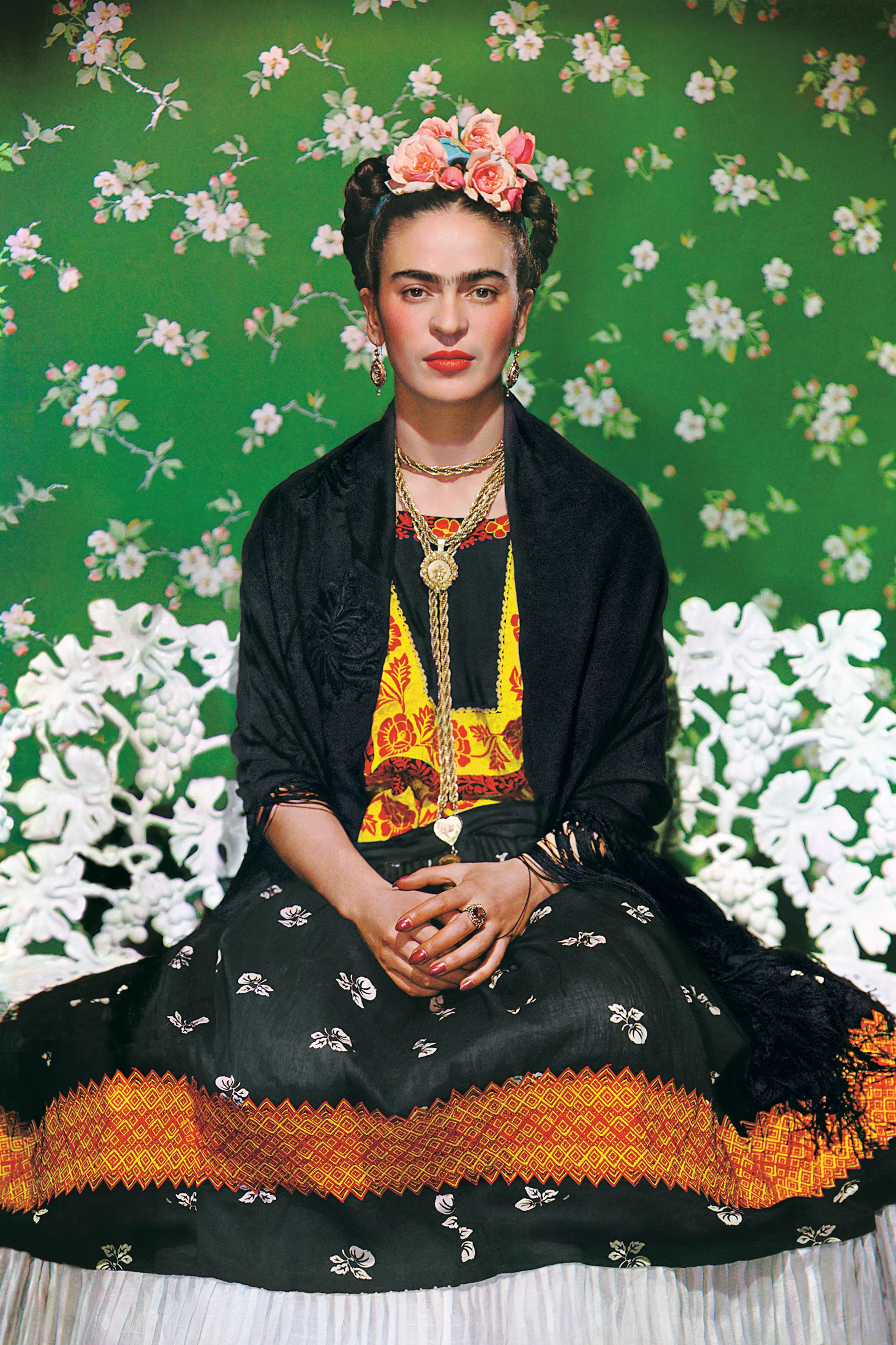Cette image montre l'artiste Frida Kahlo devant un mur vert vif, fixant l'objectif avec défi, vêtue de sa tenue traditionnelle mexicaine.