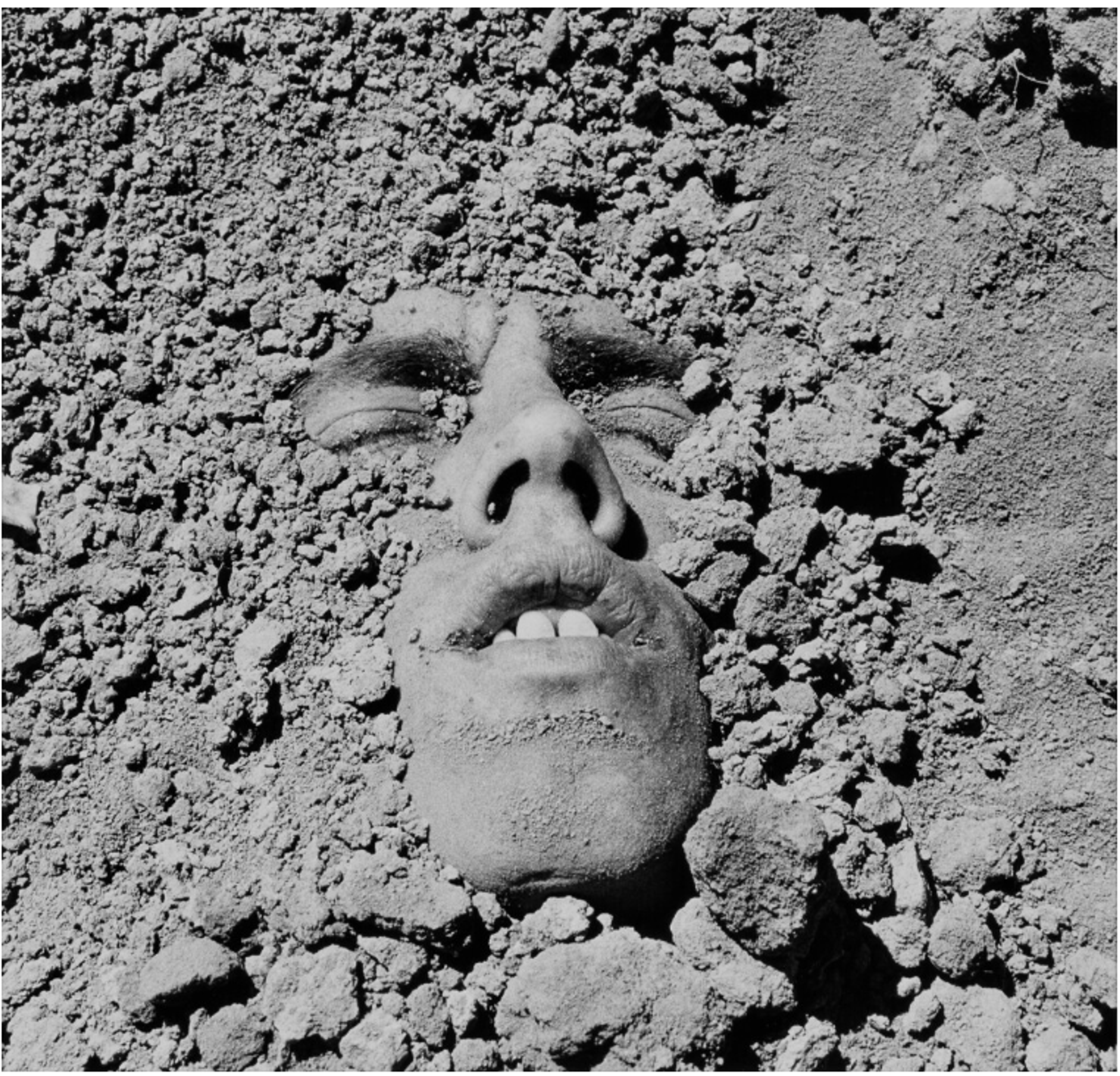 A black-and-white photograph of a man’s face half-buried in dry dirt. The eyes, nose, mouth, and chin are just above the surface. His eyes are closed in an expression of determination.