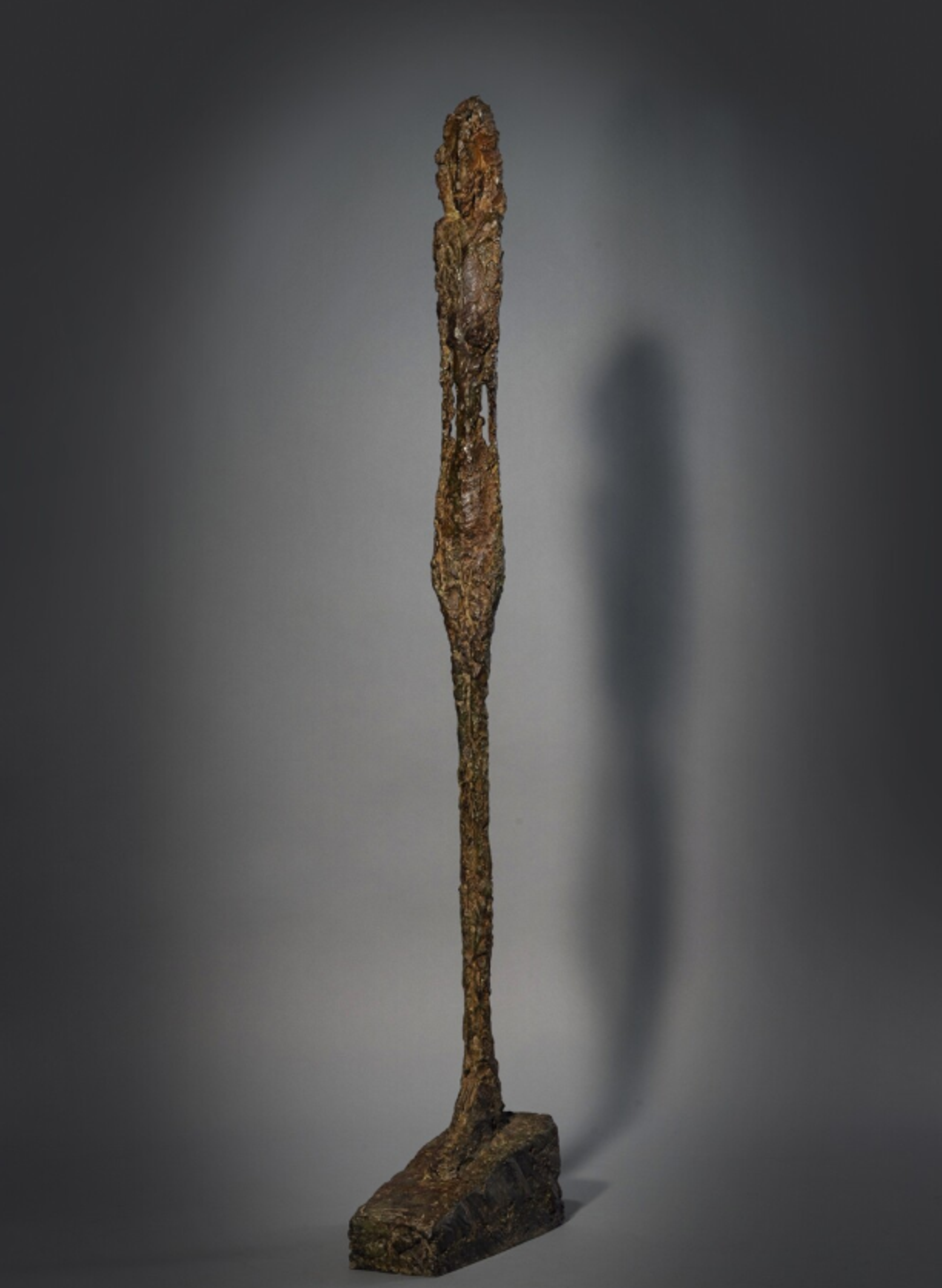 Bronze sculpture of an extremely thin, elongated standing figure with a small head. The textured surface gives the impression of rough, organic material, while the figure's impossibly slender proportions create a sense of fragility and verticality.