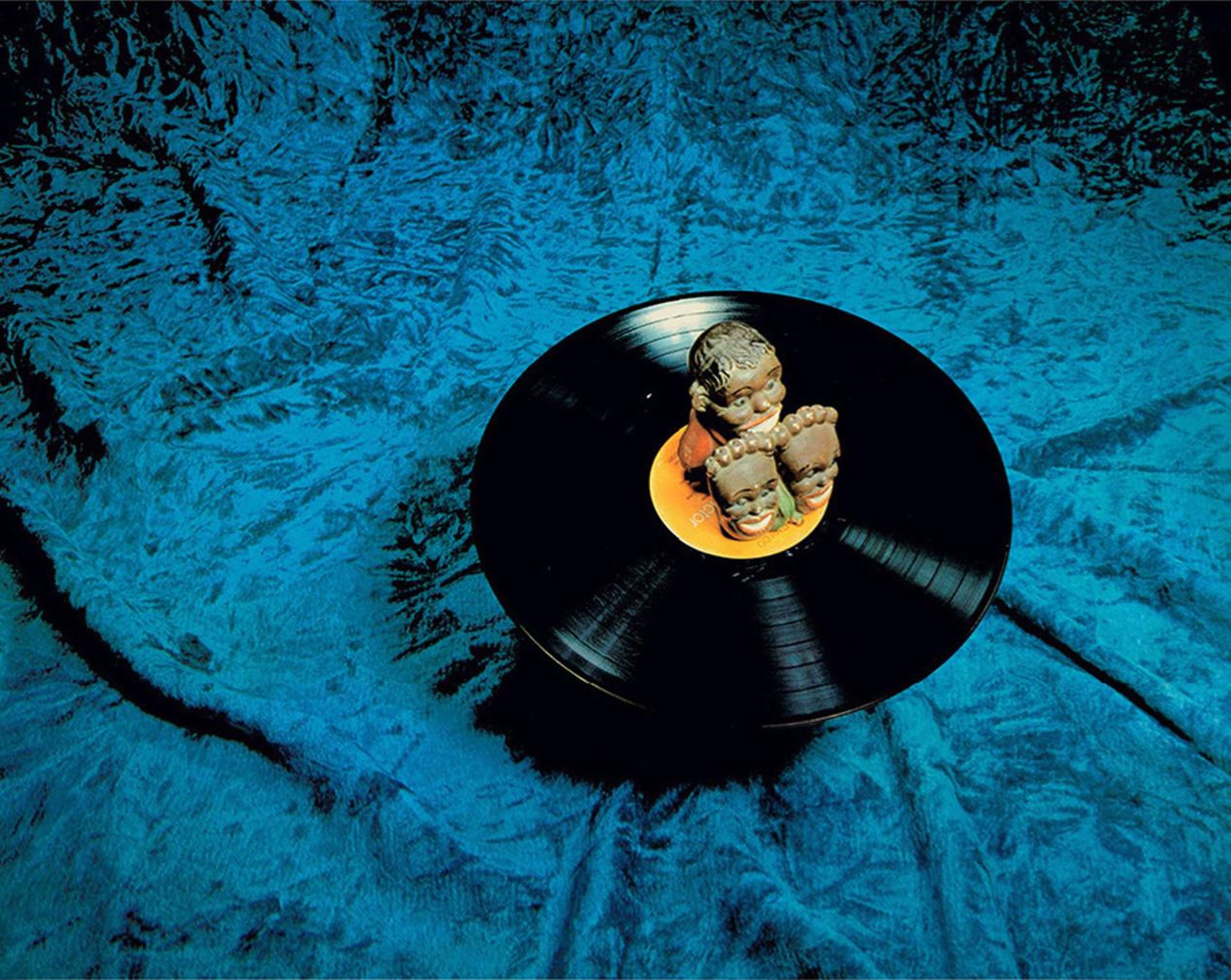 Trois têtes caricaturales en équilibre sur un disque vinyle posé sur une couverture bleue