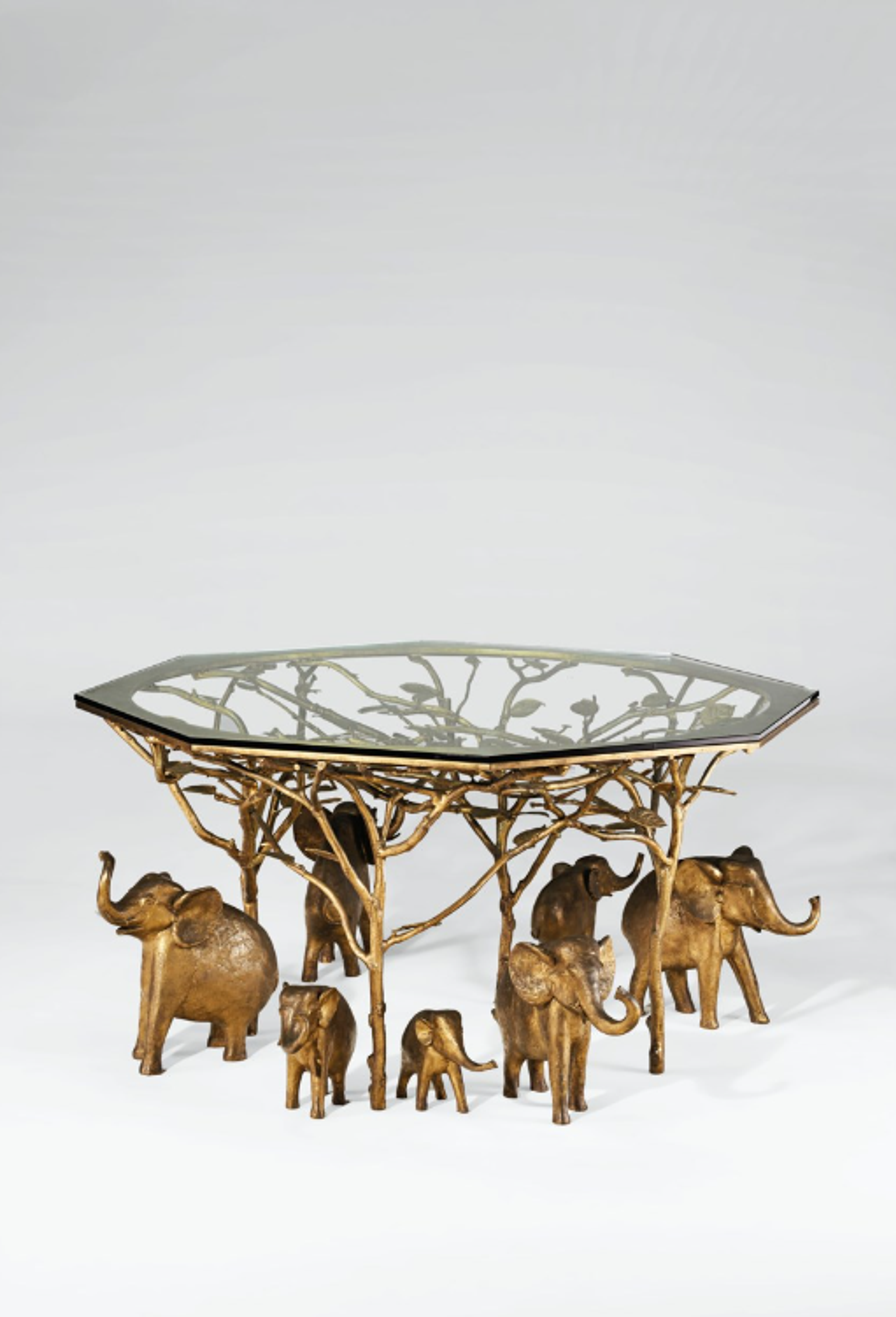 A gilded bronze sculptural piece featuring four small adult elephants and three small infant elephants beneath an intricate tree-like structure that supports a glass tabletop. Each is a separate piece, so the arrangement can be changed. The gold finish gives it an ornate, luxurious appearance.