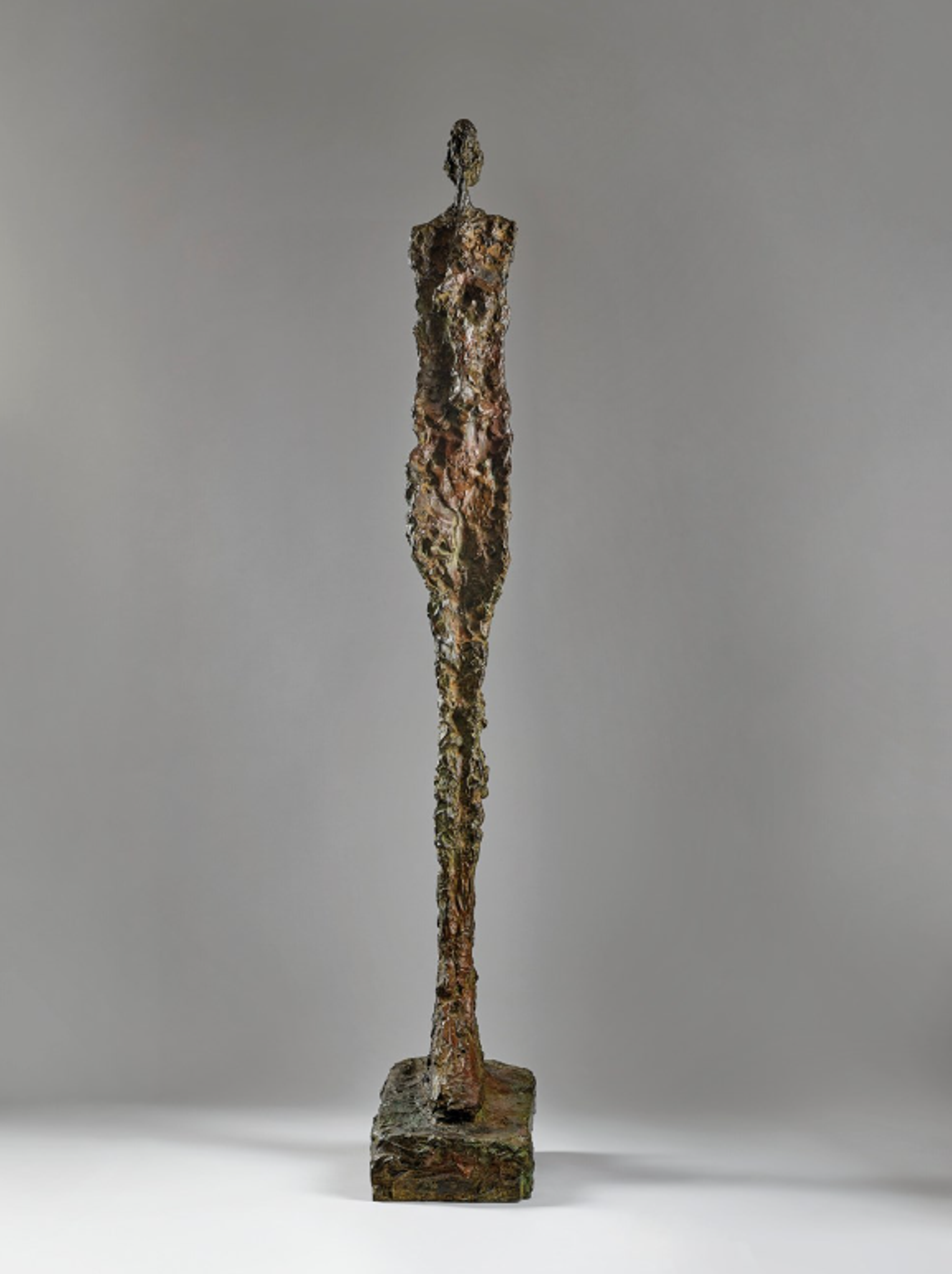 Slender bronze figure with an elongated body and simplified features, mounted on a small square base. The textured surface appears rough and weathered. The pose has arms close to the body and legs together.