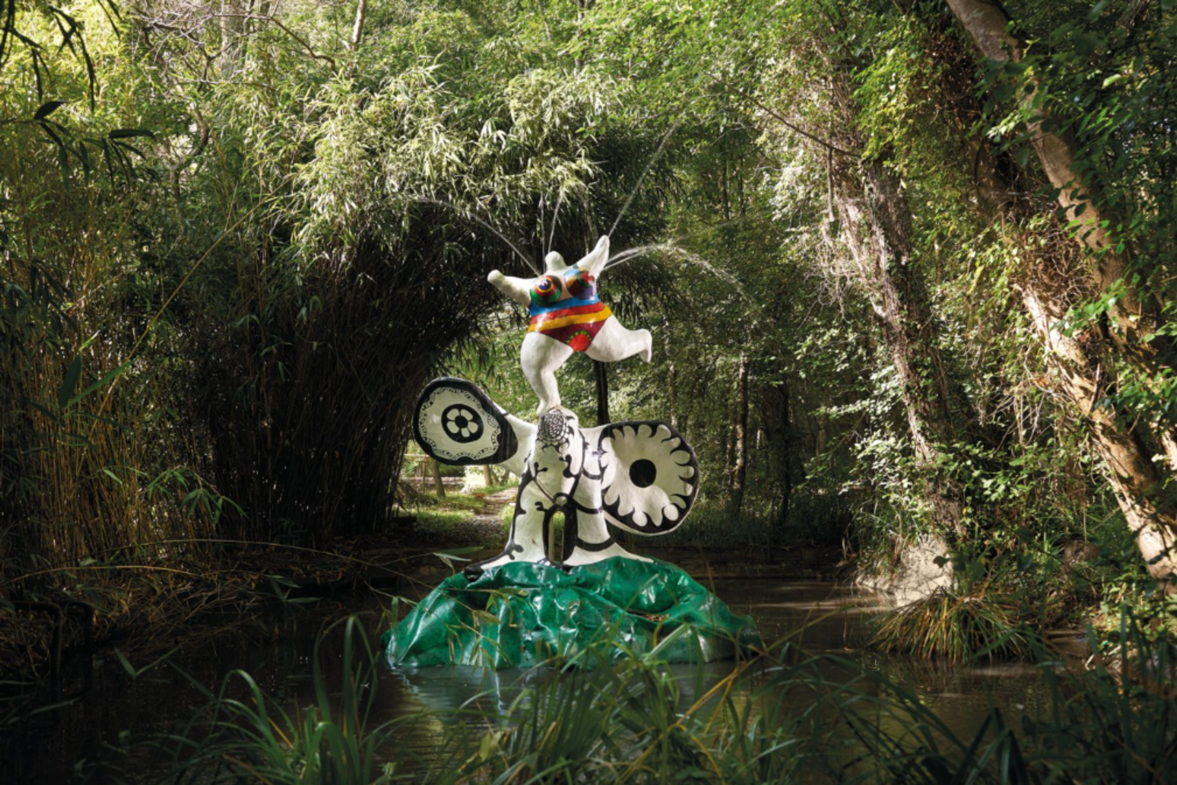 An outdoor sculpture of a white figure wearing vibrant clothes, with her arms lifted and one leg outstretched. She stands upon an abstract bird-form. The bird is black-and-white, with circular wings and four legs.