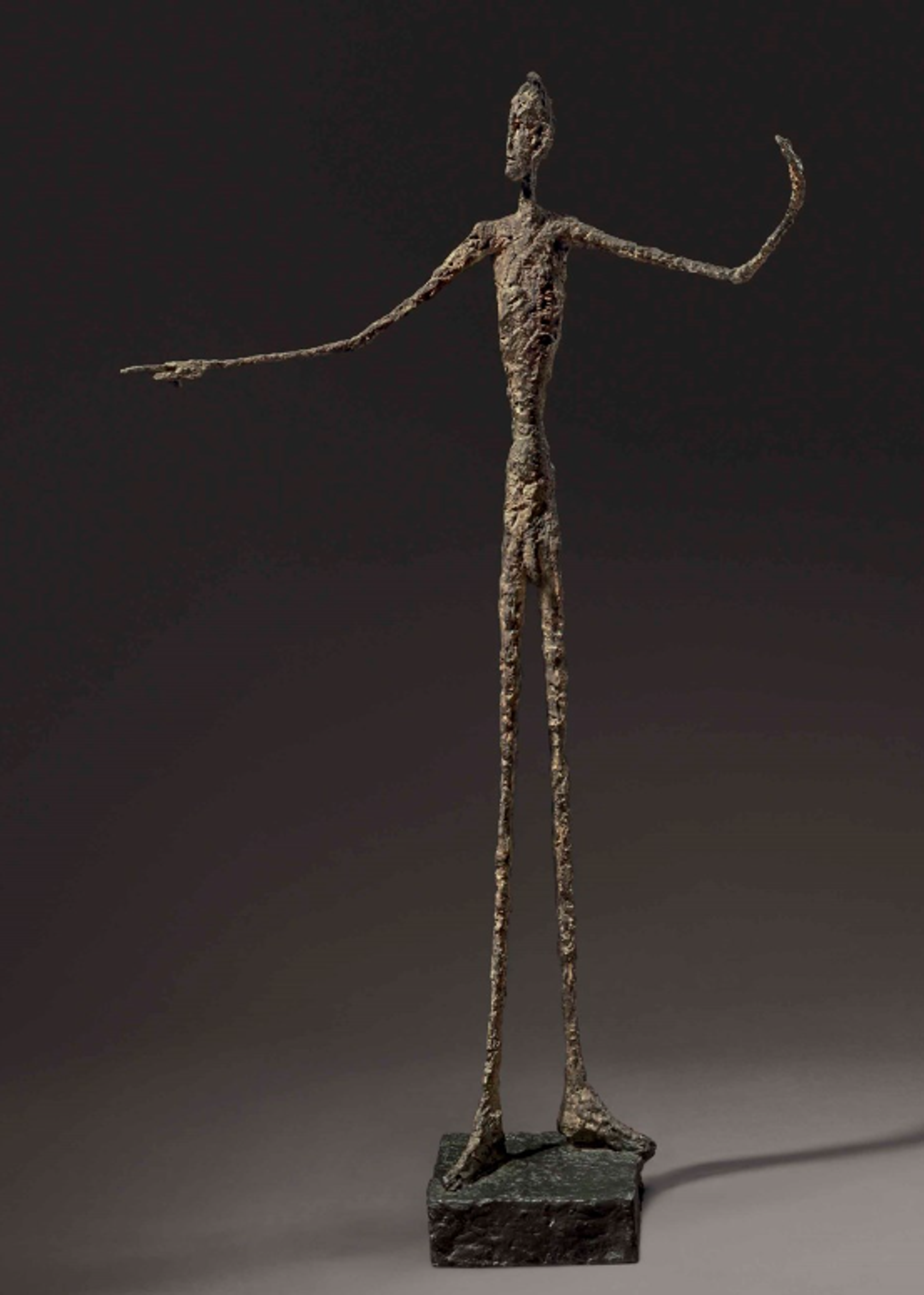 Bronze sculpture of an extremely thin, elongated figure with one arm outstretched with pointing finger and the other bent up towards the sky behind him. The figure stands on a small platform with spindly legs and arms, exhibiting a rough, textured surface and dramatic proportions.