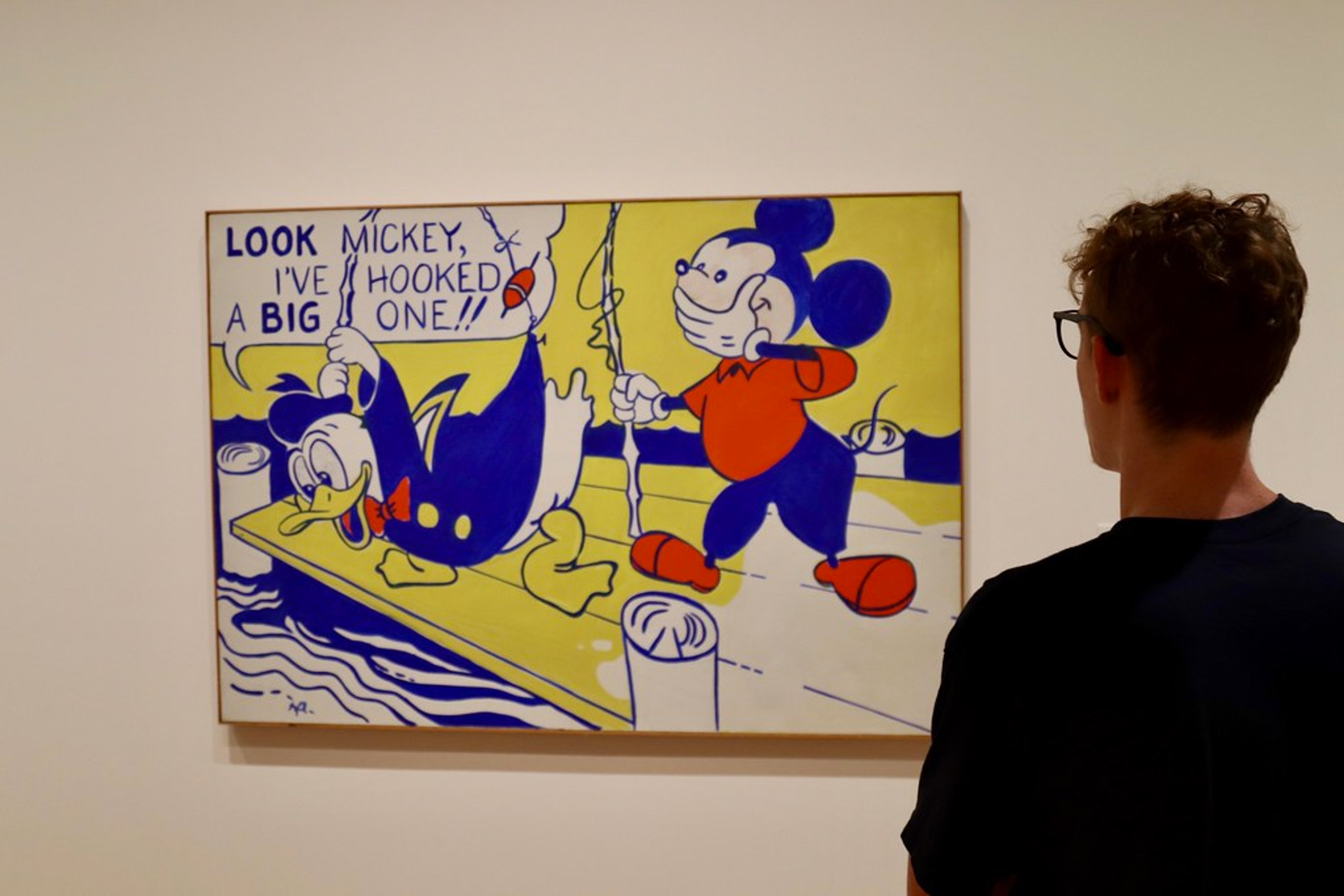 Sur cette image, un visiteur de la National Gallery de D.C. observe « Look Mickey » de Roy Lichtenstein. Les personnages de dessins animés de Disney, Donald Duck et Mickey Mouse, pêchent depuis un quai dans cette peinture horizontale. La scène et les personnages sont peints avec des aplats de jaune canari, de bleu cobalt, de rouge tomate et de blanc. À notre gauche, Donald se penche sur le bord du quai, les pieds écartés et le bec grand ouvert.