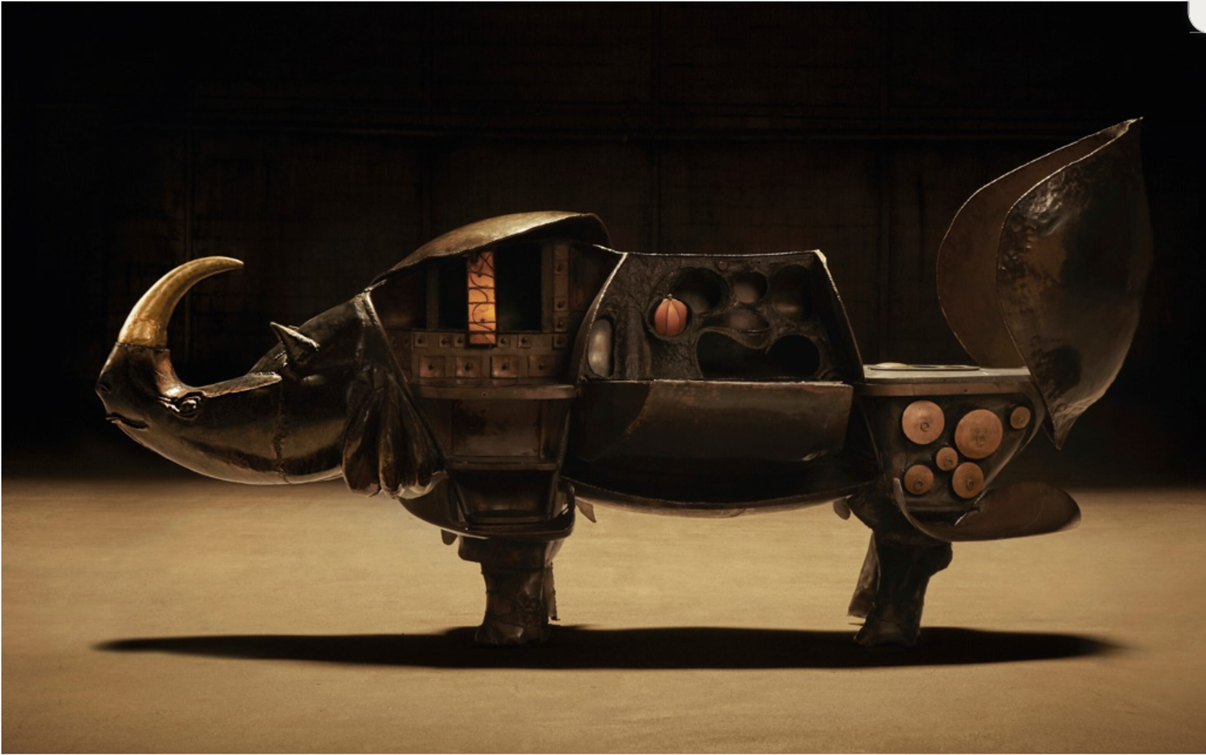 A bronze sculpture of a rhinoceros with a desk surface, shelves, and cabinets incorporated into its form. The form of the rhino appears almost robotic, with distinct sheets of metal and very sharp precise edges.