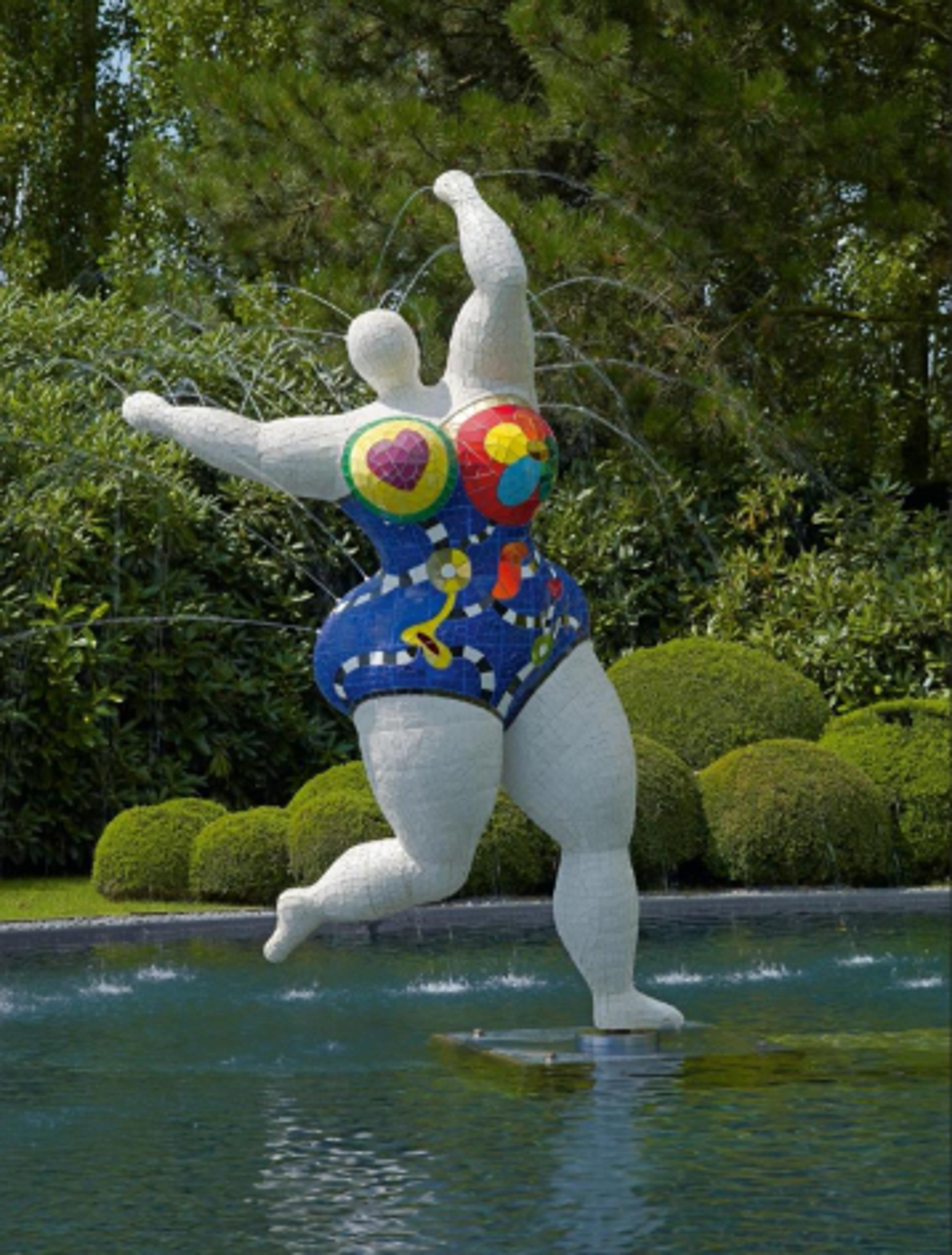 Three monumental dancing figures stand in a reflecting pool, with water spraying around them. The sculptures feature a white figure on the left, a bright yellow figure in centre, and a black figure on the right. Each figure wears a colourful, patterned swimsuit and strikes a different joyful, dancing pose.