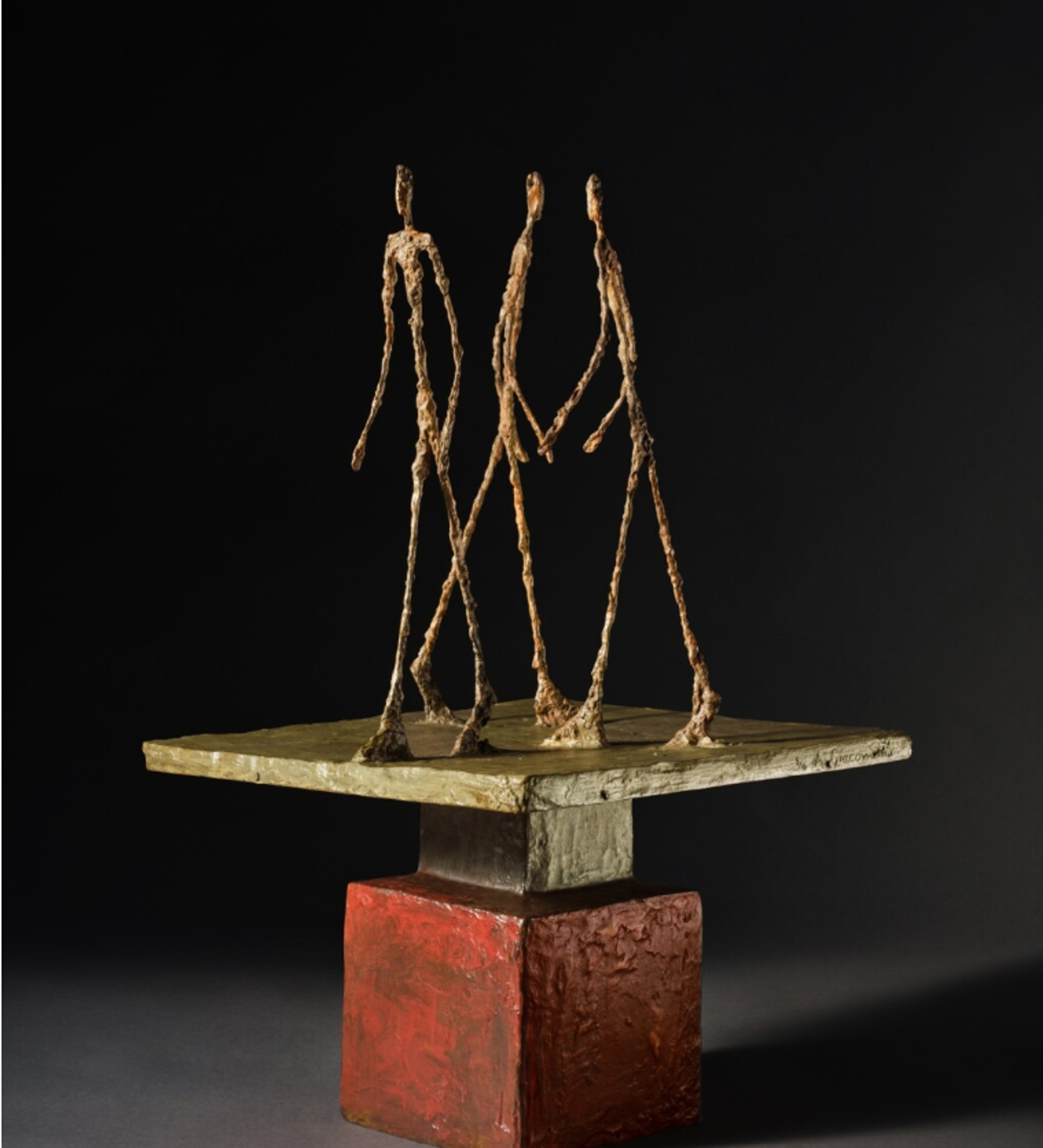 Bronze sculpture featuring three extremely thin, elongated figures arranged on a small square platform. The spindly figures appear to be in mid-stride, with long, stick-like limbs anchored to a brass-coloured base on top of a square pedestal.