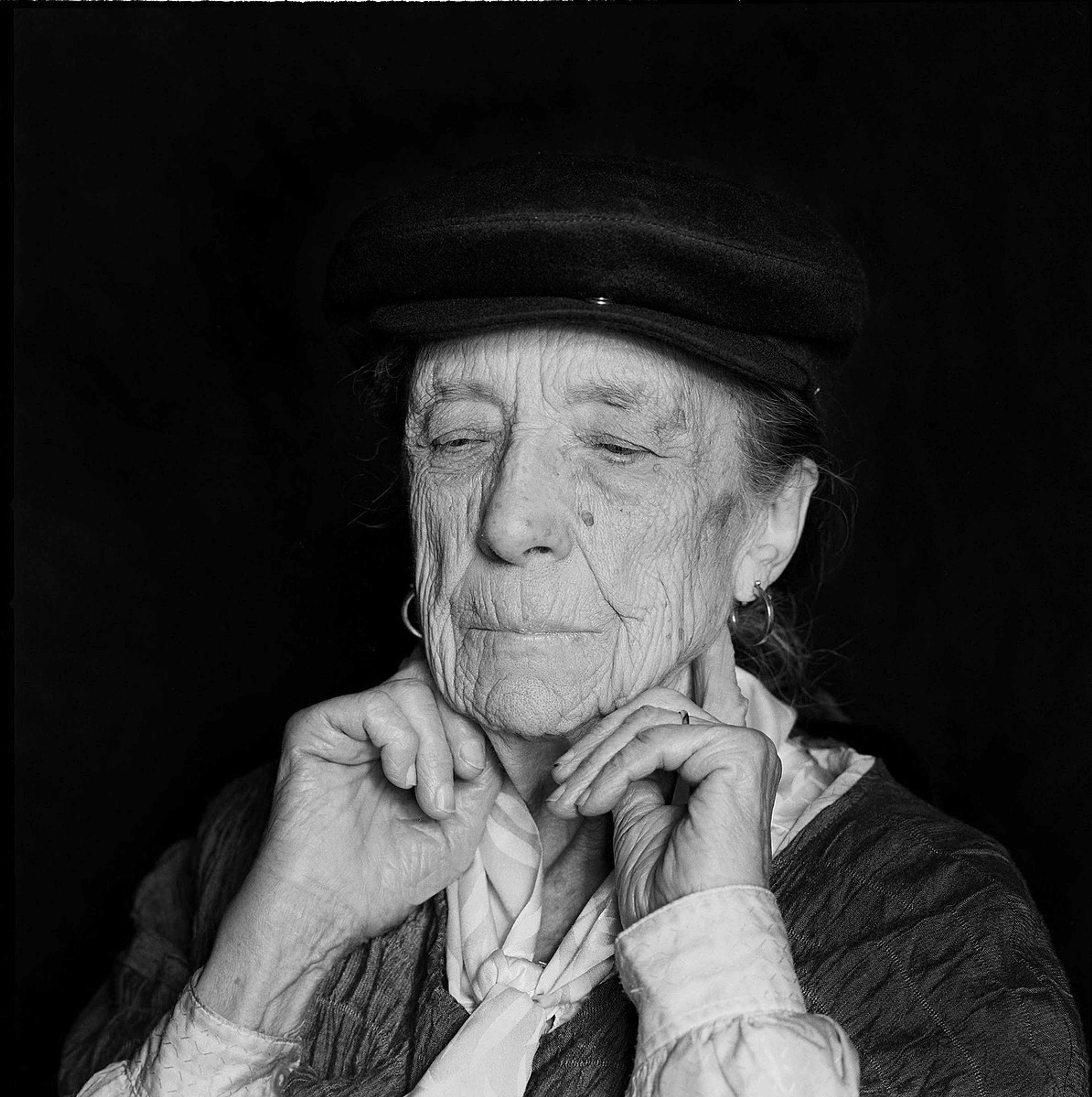 Portrait en noir et blanc d'une femme âgée se tenant le visage, vêtue d'un chapeau et d'une blouse.