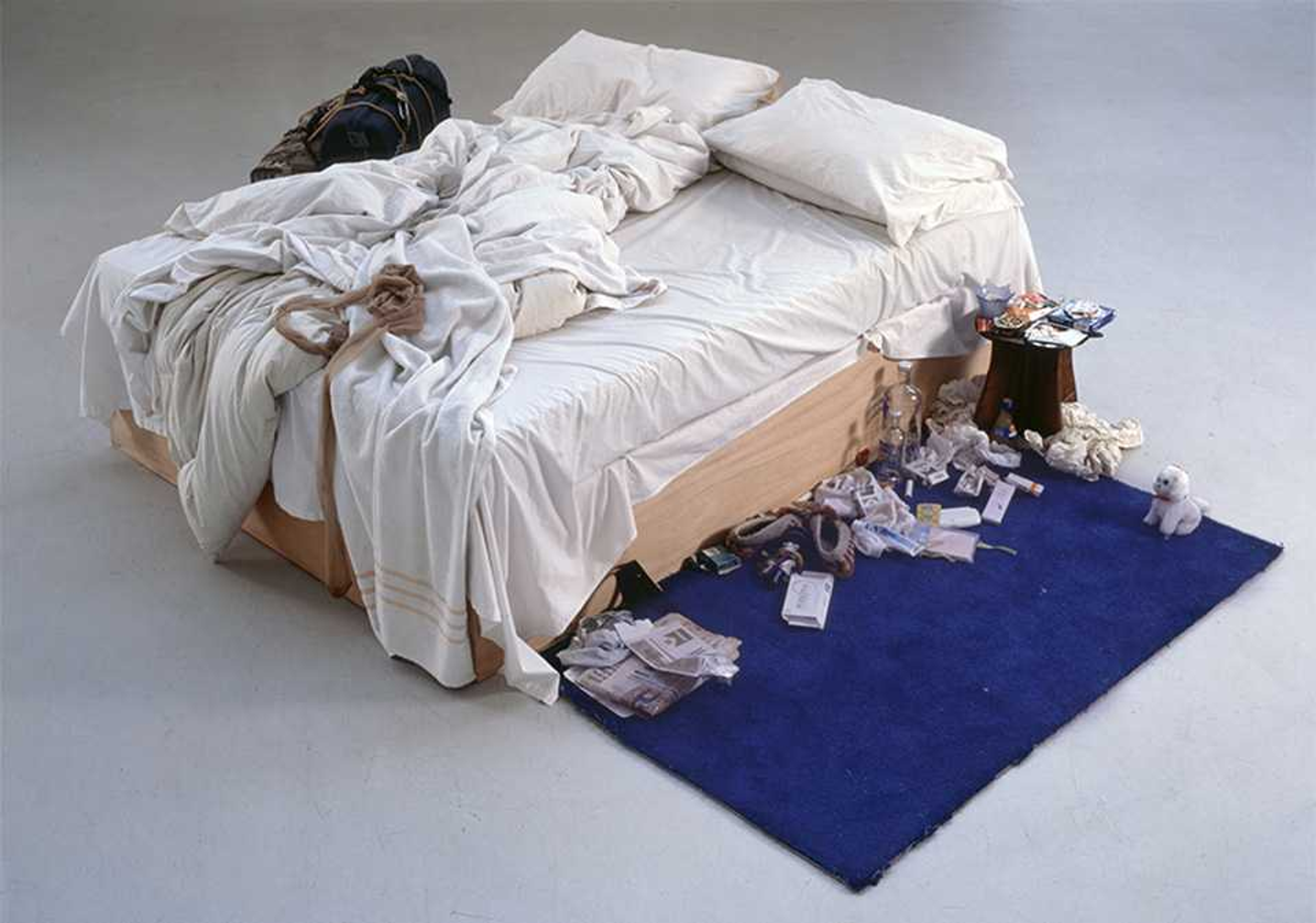 Photographie de « My Bed » de Tracey Emin. Un lit défait avec une literie en boule, à côté d'un tapis bleu jonché d'objets divers et personnels.