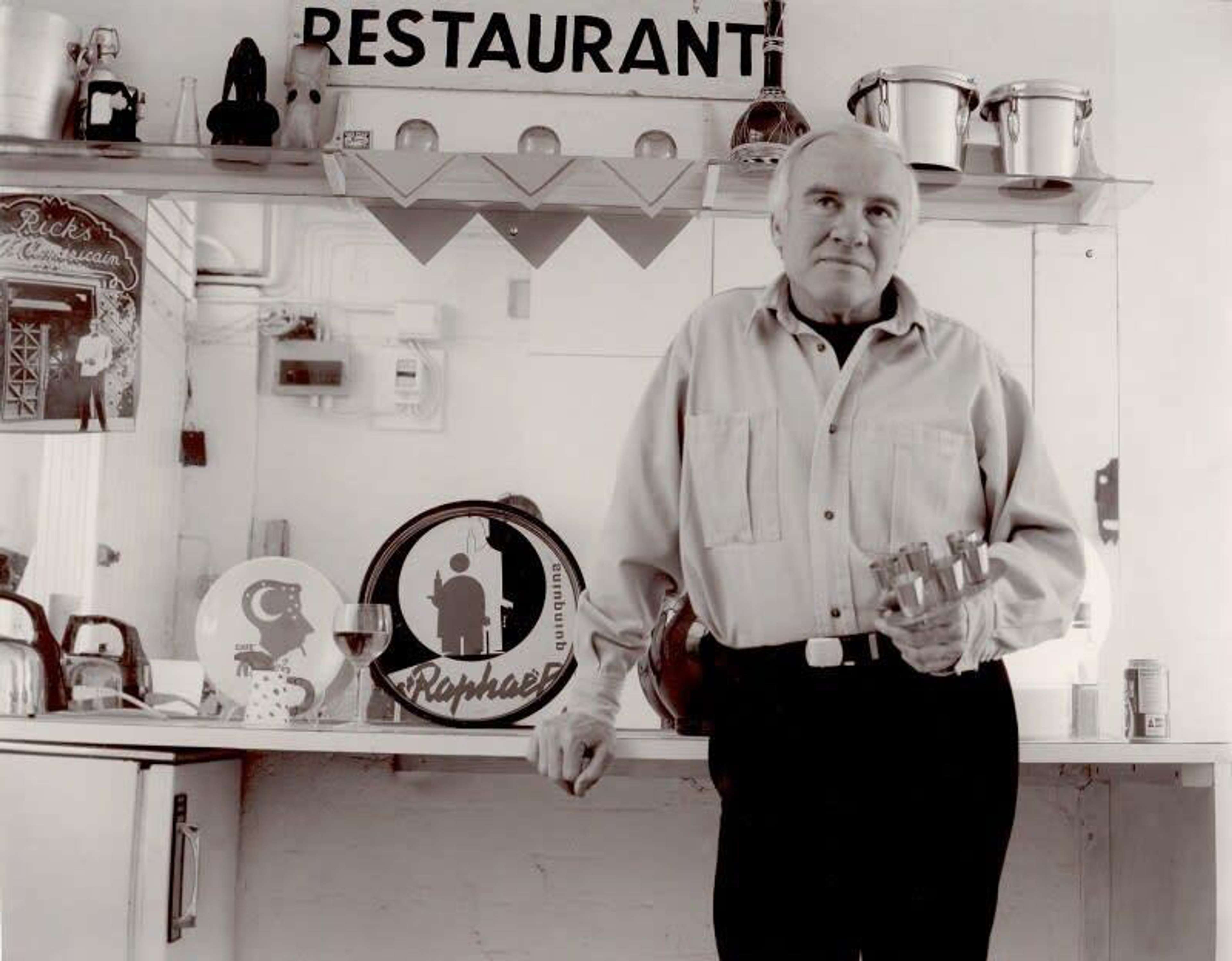 Une image en noir et blanc de l'artiste Patrick Caulfield, debout devant une cuisine.
